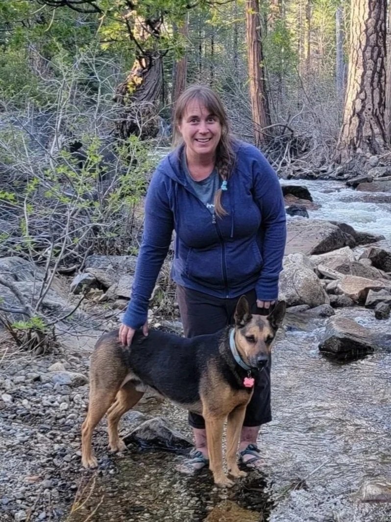 A woman in a blue jacket standing in a shallow creek with a German Shepherd dog, surrounded by trees and rocks in a forested landscape.