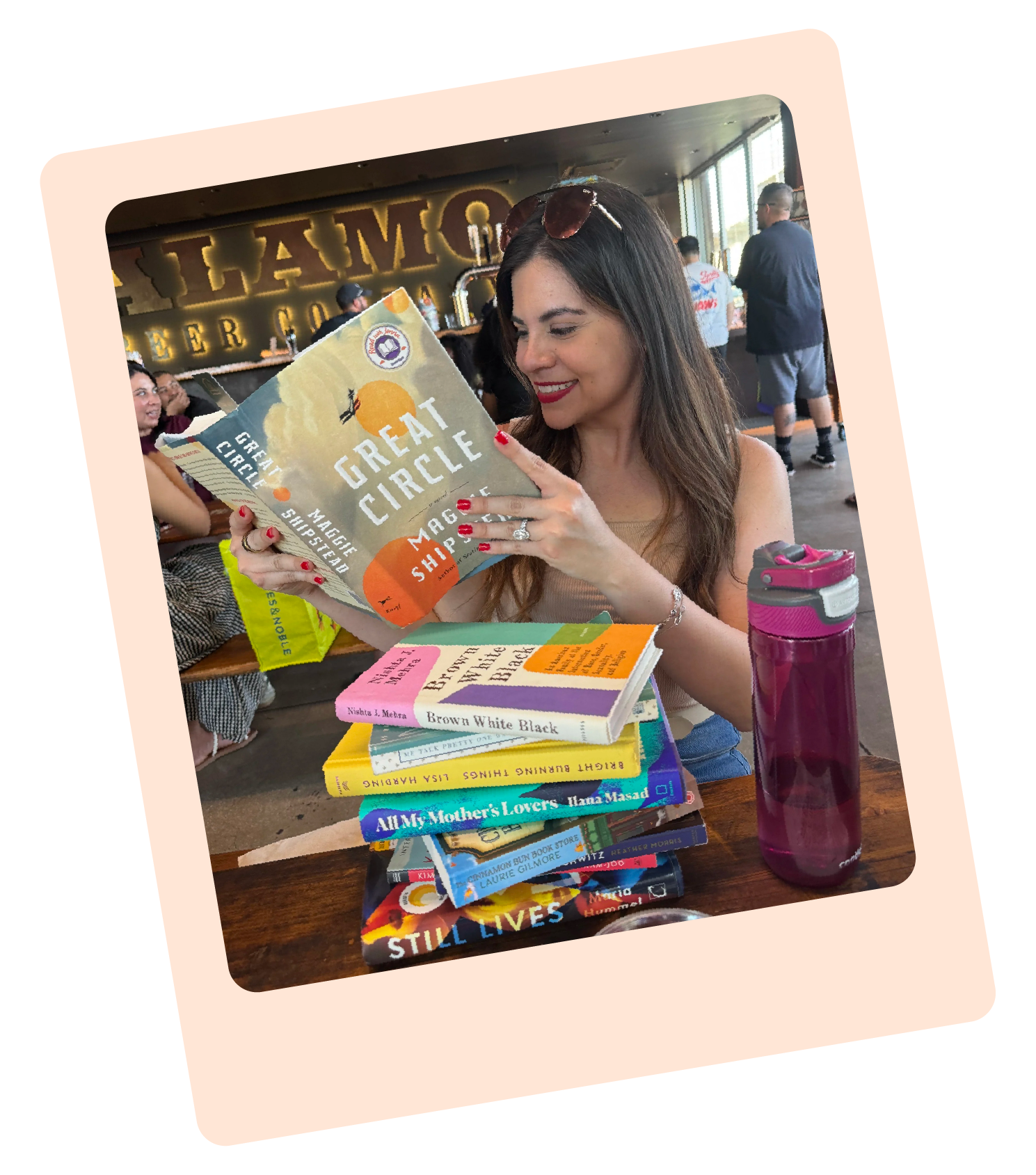 A polaroid style photo showing Luana, founder of Thread Studio, sat reading a book. On the table in front of her is a stack of colorful books and a pink water bottle.