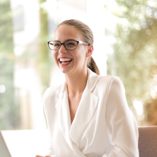 A woman with glasses and a white blazer smiling and laughing indoors with sunlight and trees visible through window behind her.