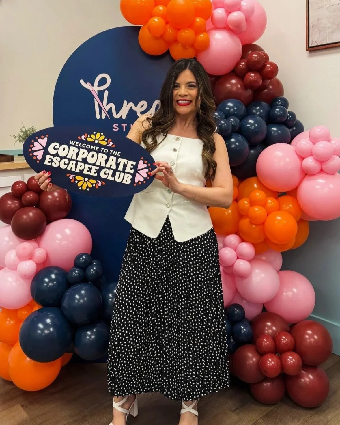Luana, founder of Thread Studio, smiling at the camera holding a sign that reads 'Welcome to the Corporate Escapee Club' in front of a colorful balloon backdrop, in San Antonio, Texas.