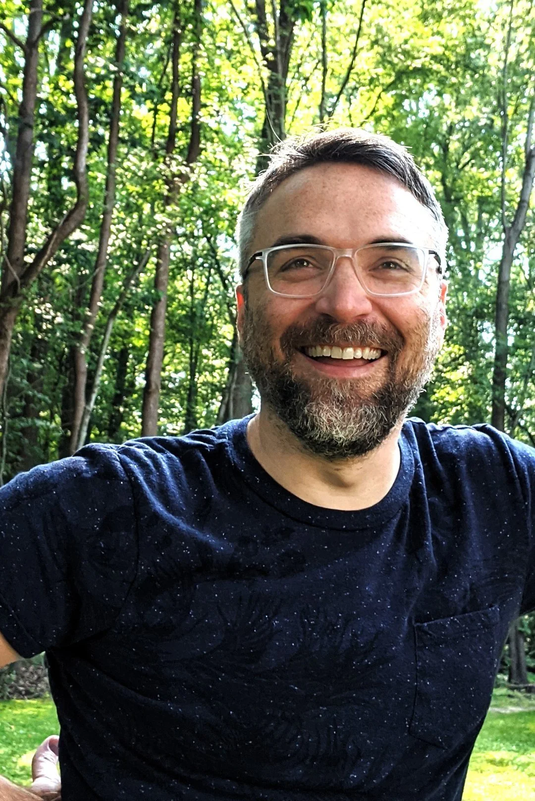 A smiling man with glasses and a beard outdoors in a wooded area with green trees and sunlight filtering through.