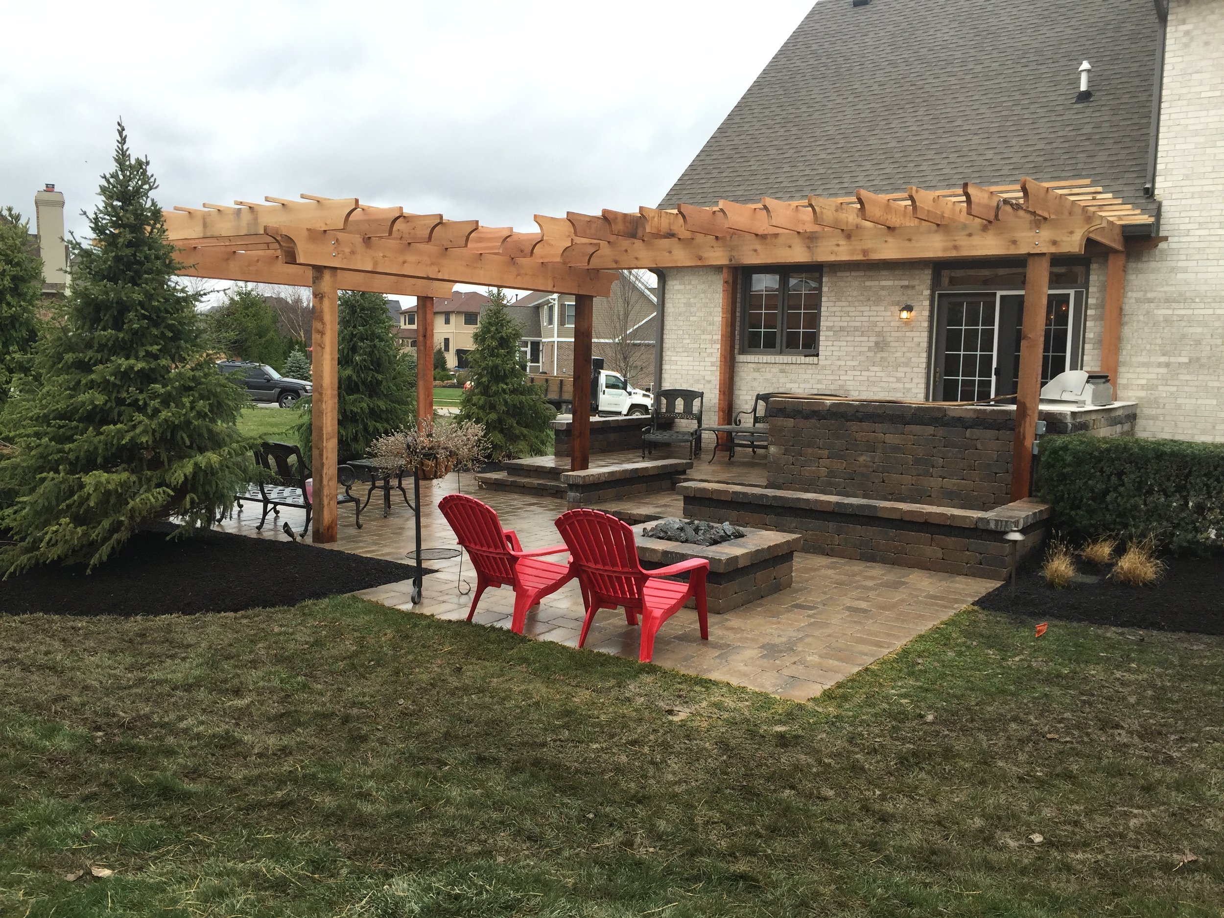 Backyard patio with brick fireplace, wooden pergola, lawn chairs, and potted plants, adjacent to a brick house.