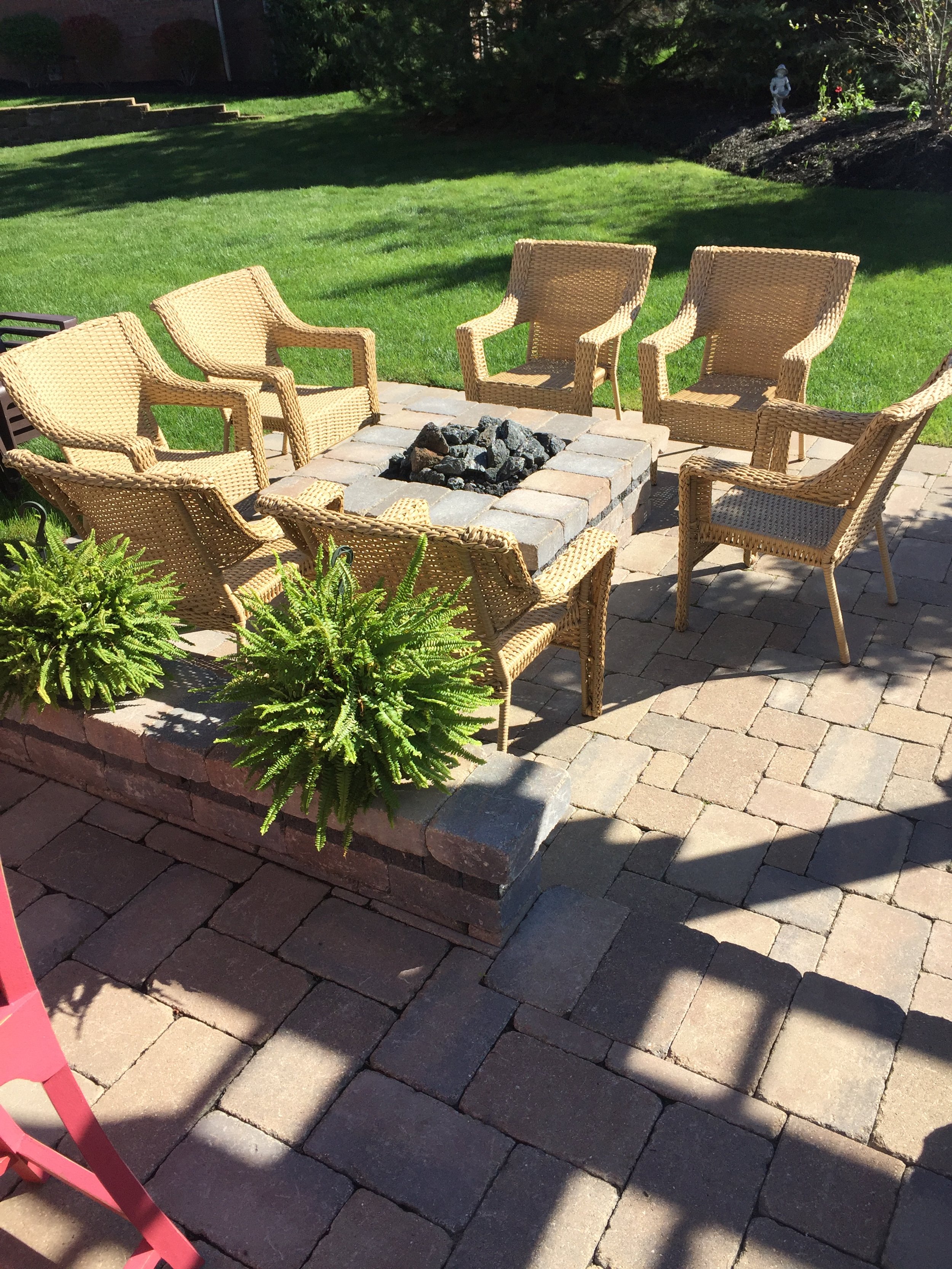 Outdoor patio with six wicker chairs arranged around a square fire pit on a brick paver surface, with a grassy yard and garden in the background.