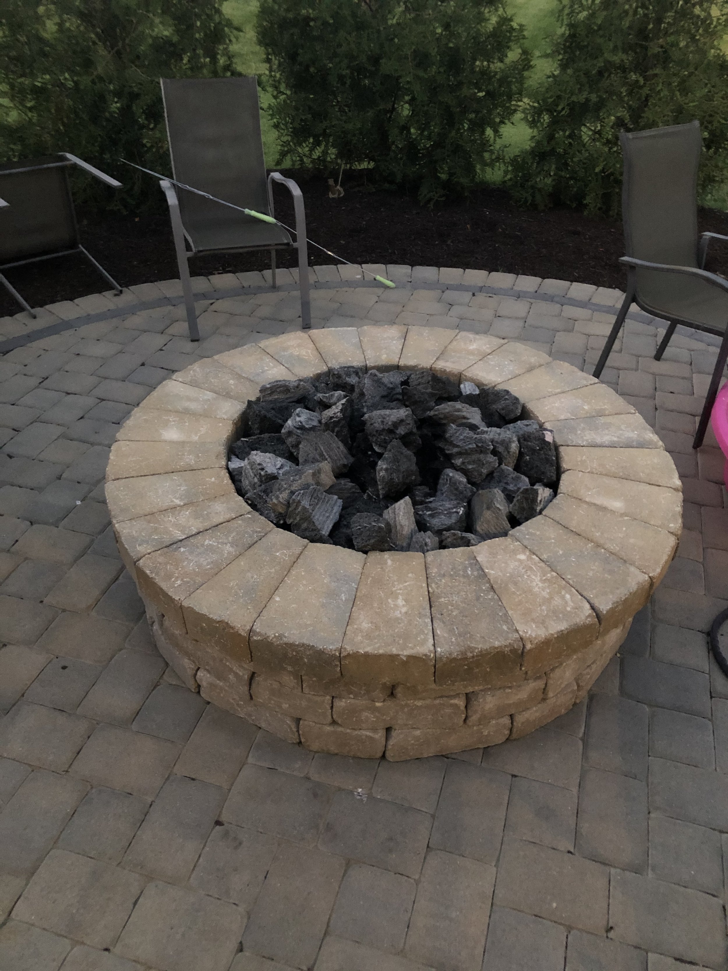 A circular fire pit made of tan bricks with black charcoal inside, surrounded by a paver patio and outdoor chairs, with greenery in the background.