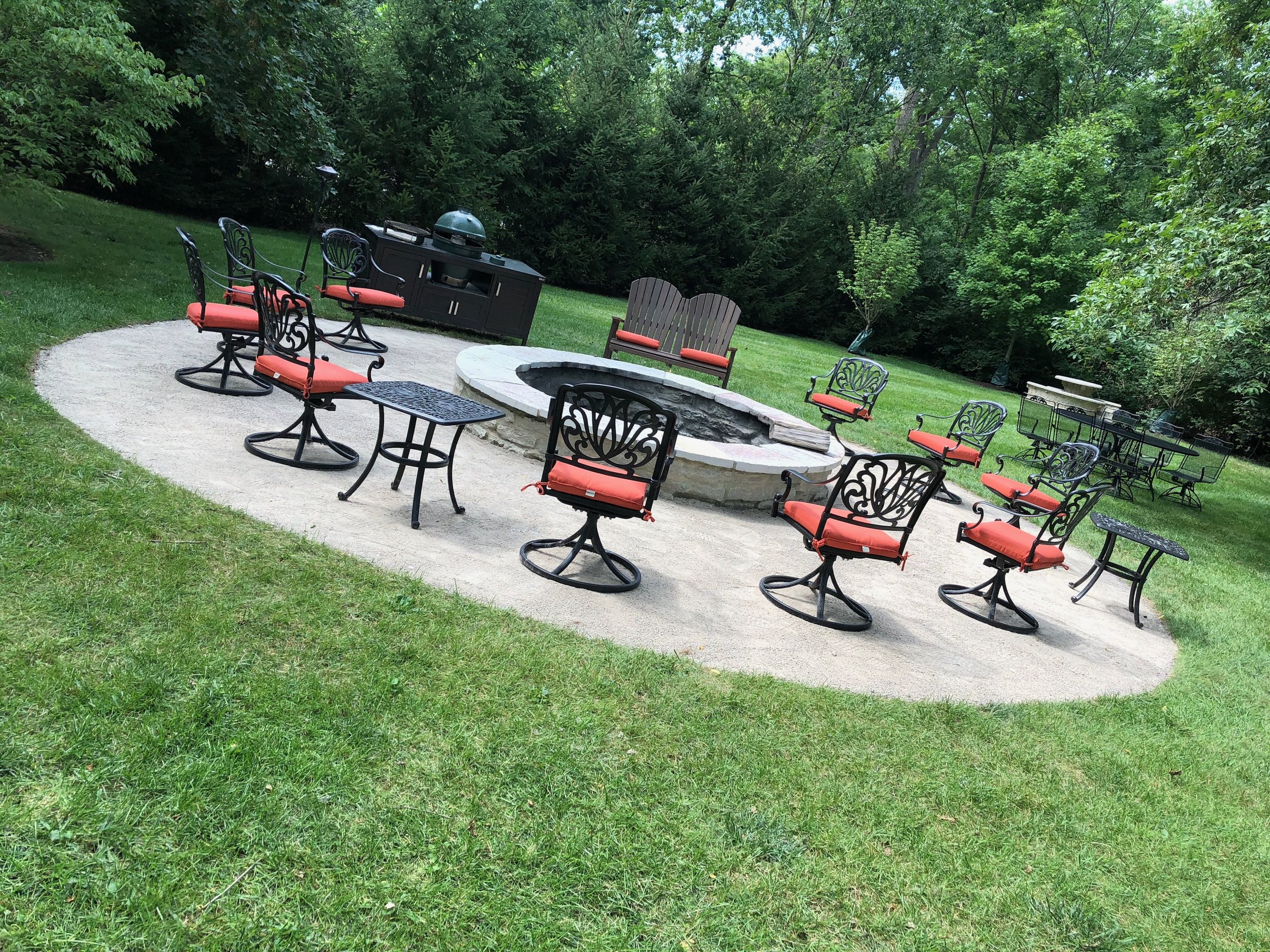 Outdoor backyard patio with multiple black wrought iron chairs with red cushions around a fire pit, a stone barbecue grill, and green trees in the background.