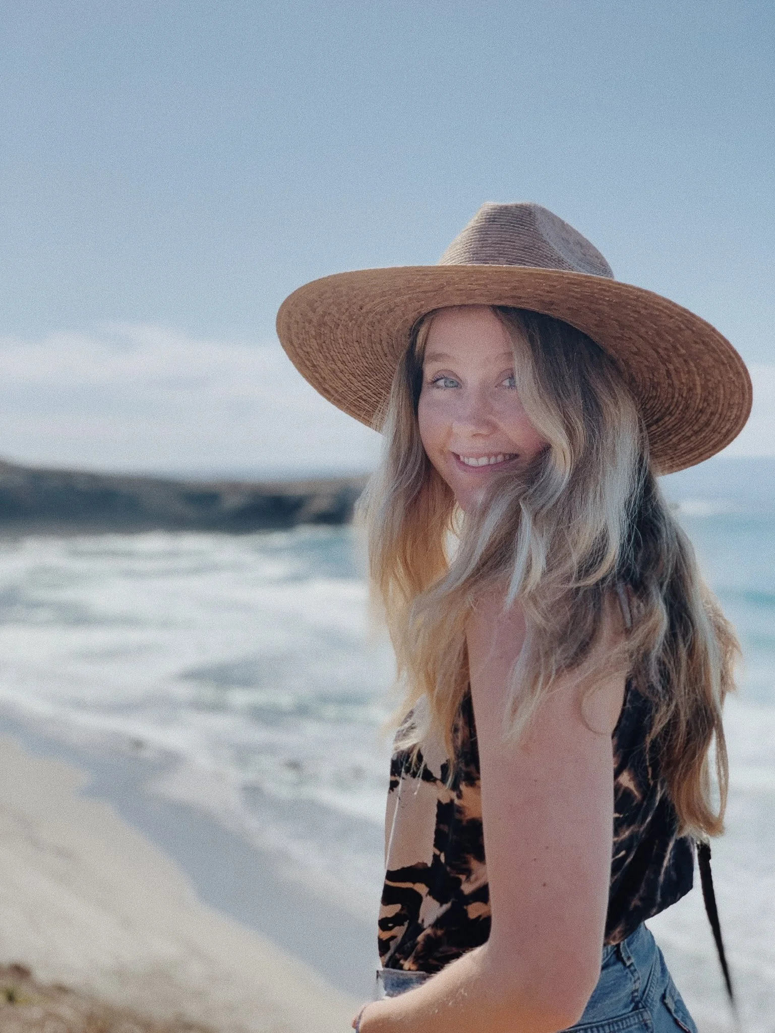 Arianna Ross, a life coach in LA, smiling wearing a wide-brimmed straw hat and a sleeveless animal print top, standing on a beach with ocean waves in the background.