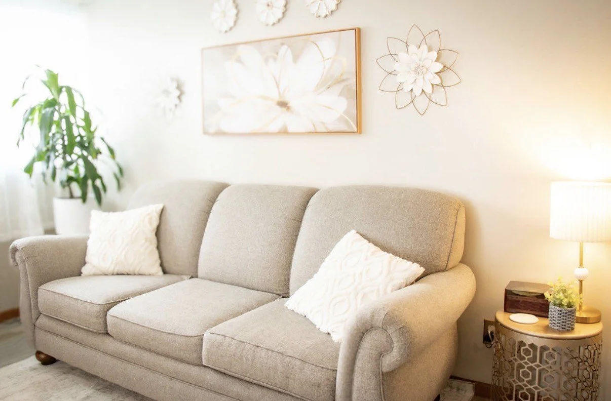 A beige sofa with two white textured pillows, a side table with a lamp, a potted plant, and wall decorations in a well-lit living room.
