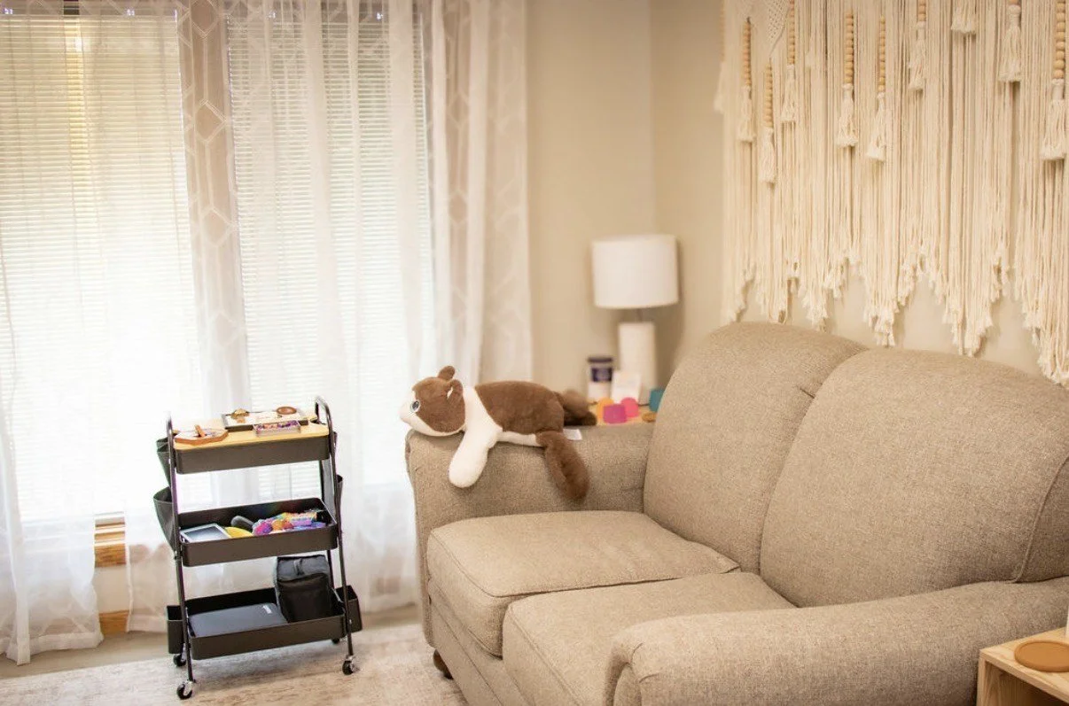 Living room with beige sofa, stuffed horse toy on the armrest, side table with colorful toys, standing lamp, and sheer curtains covering windows.