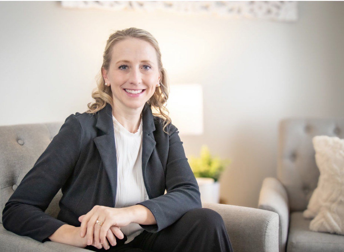 A woman wearing a black blazer and white blouse sitting on a gray couch, smiling at the camera in a well-lit living room.