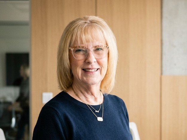 A smiling middle-aged woman with blonde hair and glasses who greets clients warmly, wearing a navy sweater, standing indoors against a wooden-paneled wall.
