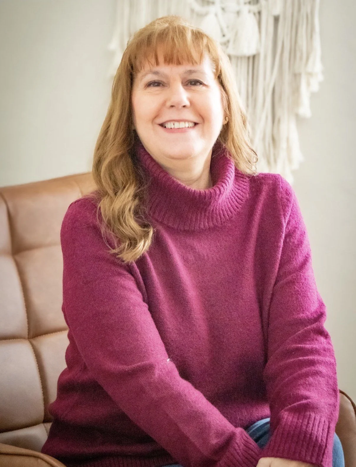 A woman with shoulder-length red hair and fair skin, smiling while sitting on a leather chair, wearing a magenta turtleneck sweater, with a cream-colored woven wall hanging in the background.
