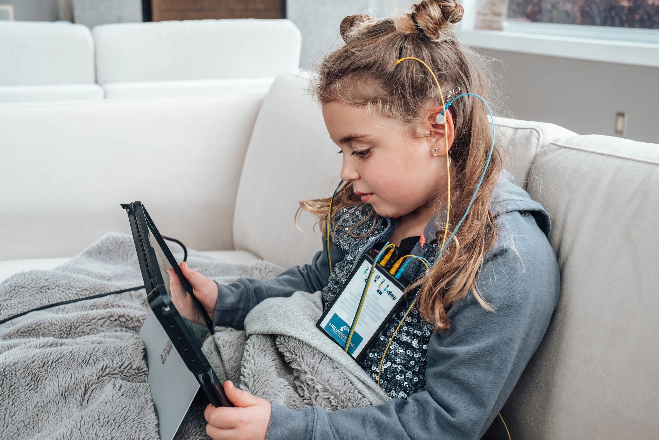 A young girl with long hair and earrings sitting on a beige couch, using a tablet with a portable neurostimulation device attached to her chest, connected by yellow and blue wires, wrapped in a grey blanket.