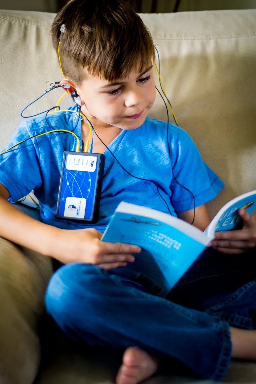 A young boy sitting on a beige couch, wearing a blue T-shirt and jeans, reading a book while connected to a mental health or brain activity device with wires attached to his head and a small screen hanging around his neck.