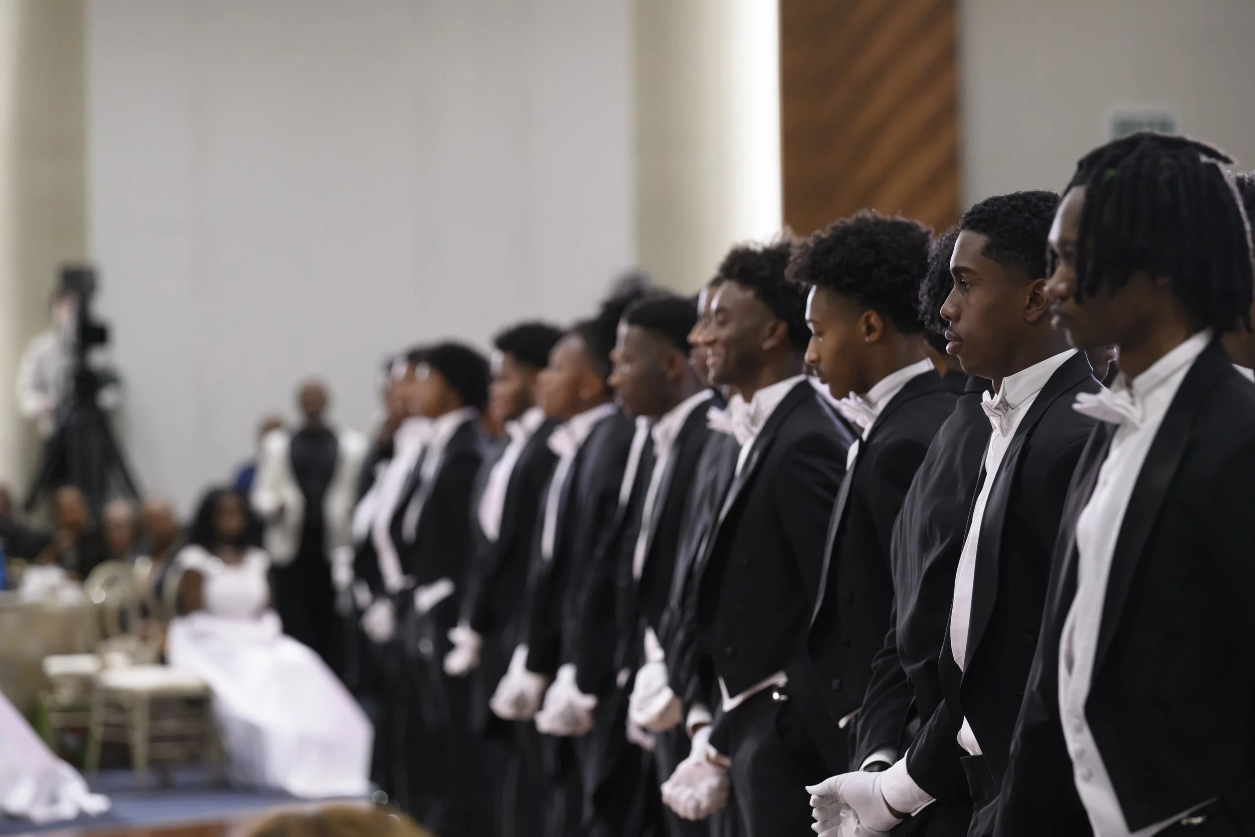 Group of young men in tuxedos with white gloves standing in a line at a formal event