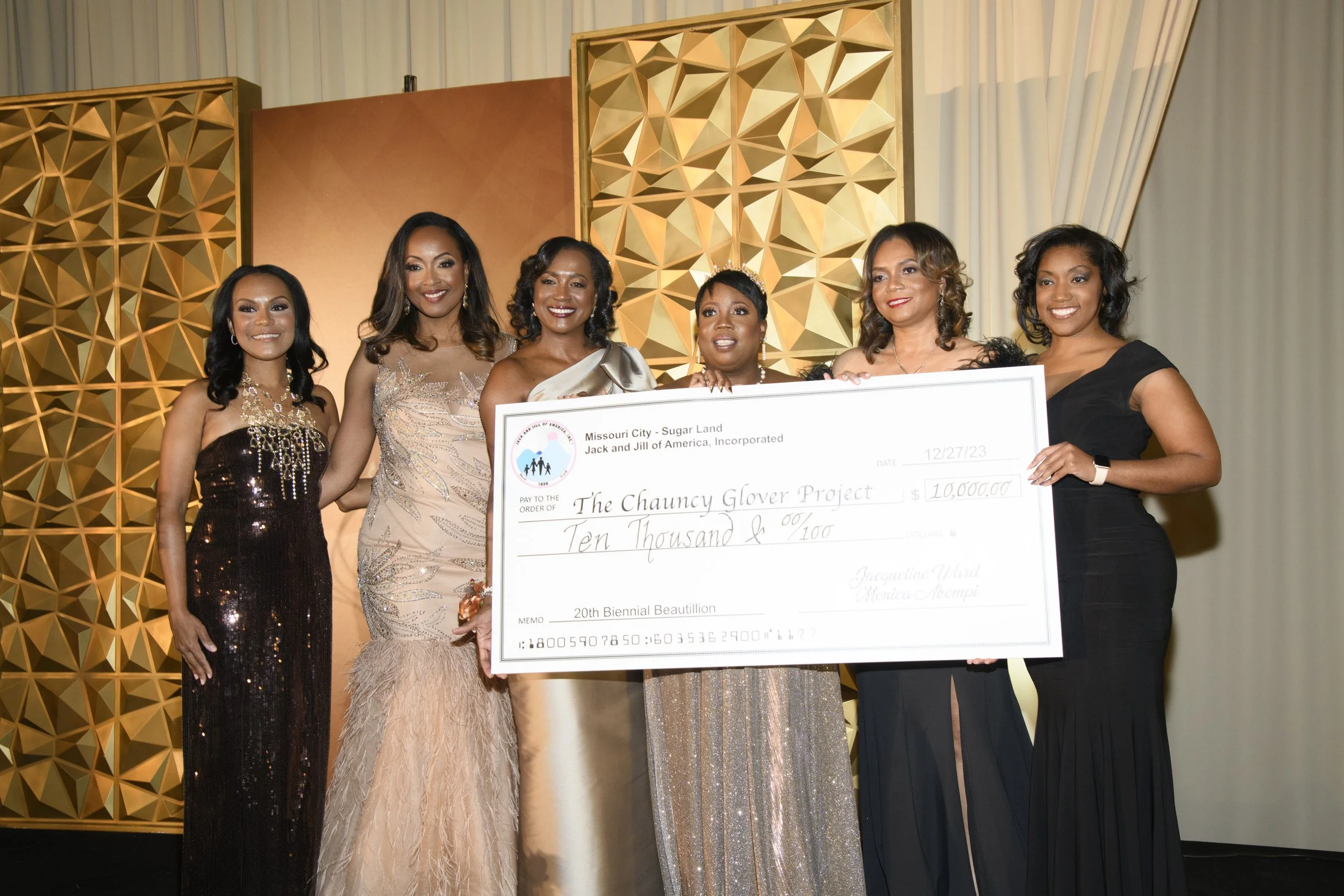 Six women in evening dresses standing together on stage holding a large check made out to The Chauncy Glover Project for ten thousand dollars, with a gold decorative backdrop behind them.