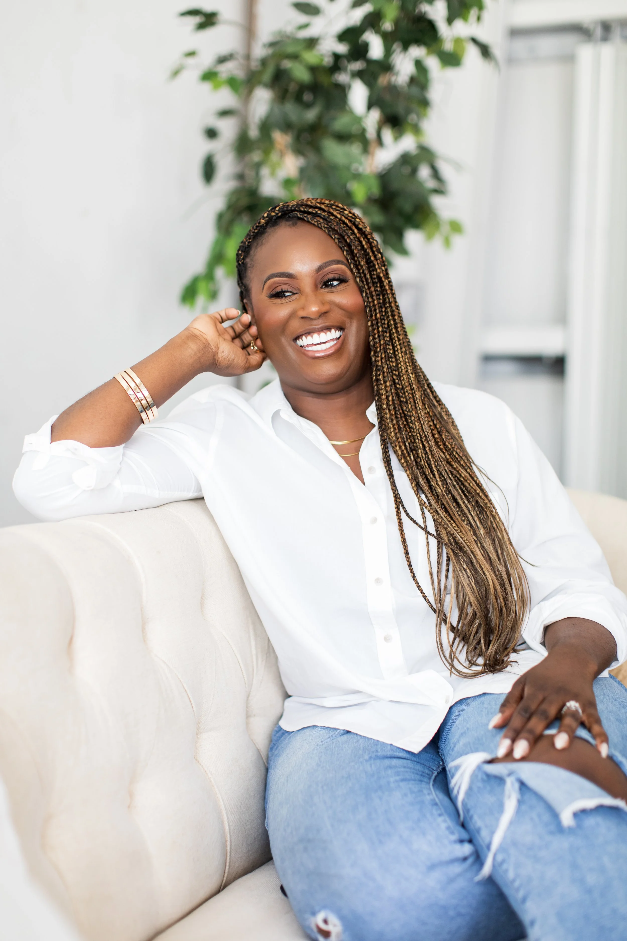 A smiling woman with long braided hair sitting on a beige sofa, wearing a white shirt and ripped jeans, with a green plant in the background.