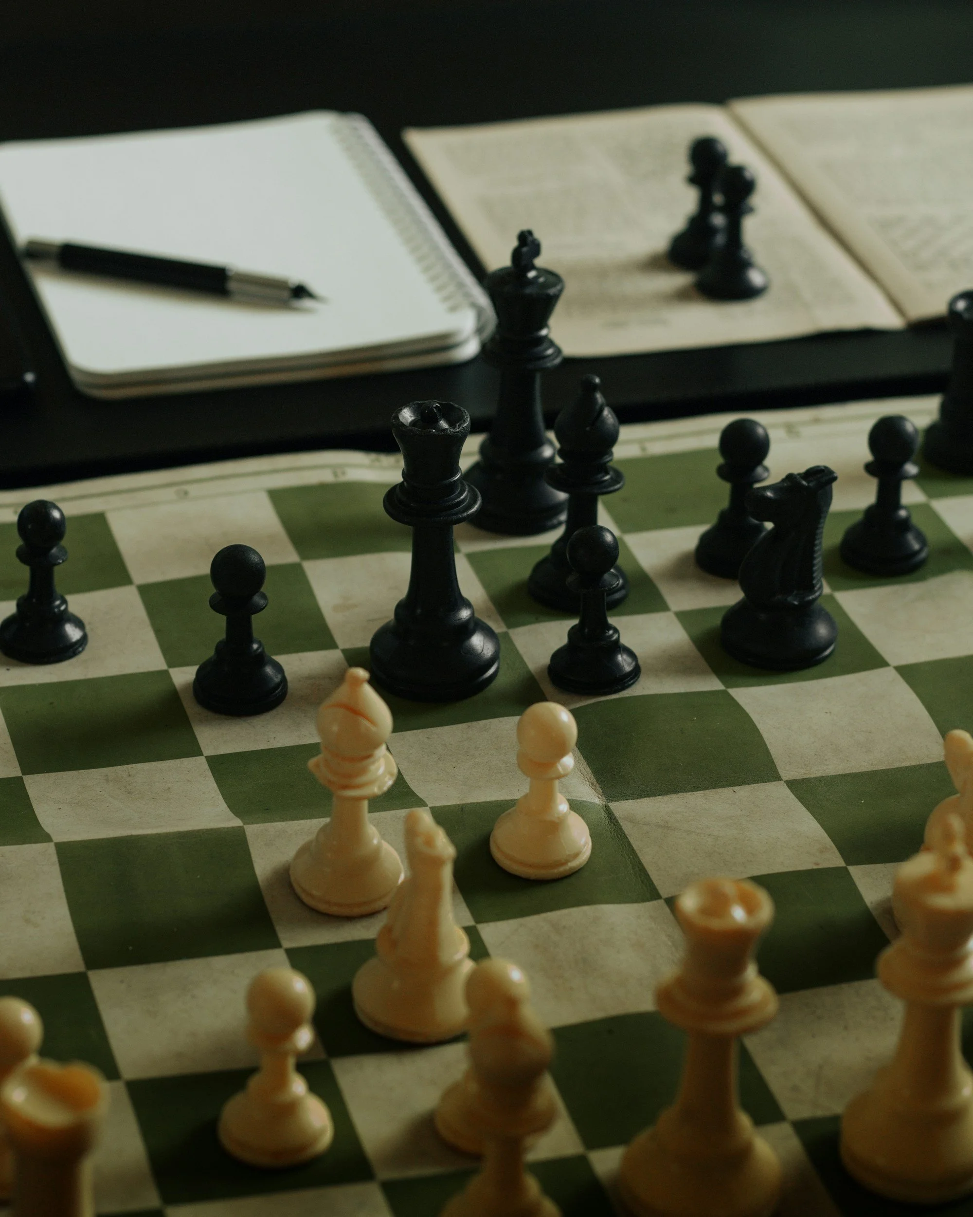 A chessboard with black and white pieces set up, with notebooks and a pen on a dark surface in the background.