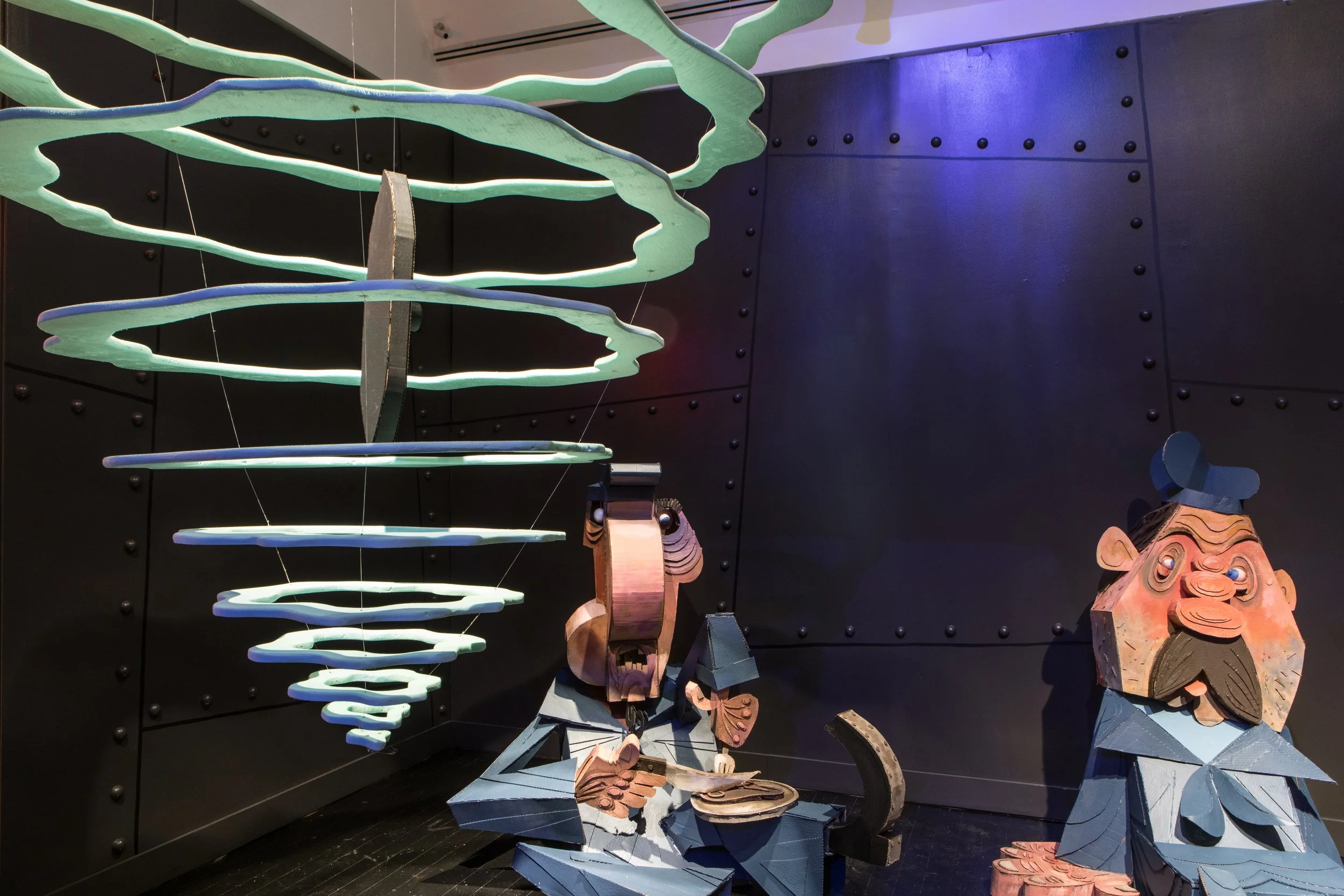 Colorful abstract sculptures of two characters, one with a mustache and hat, and another with a large head, set against a dark wall with colorful lighting.