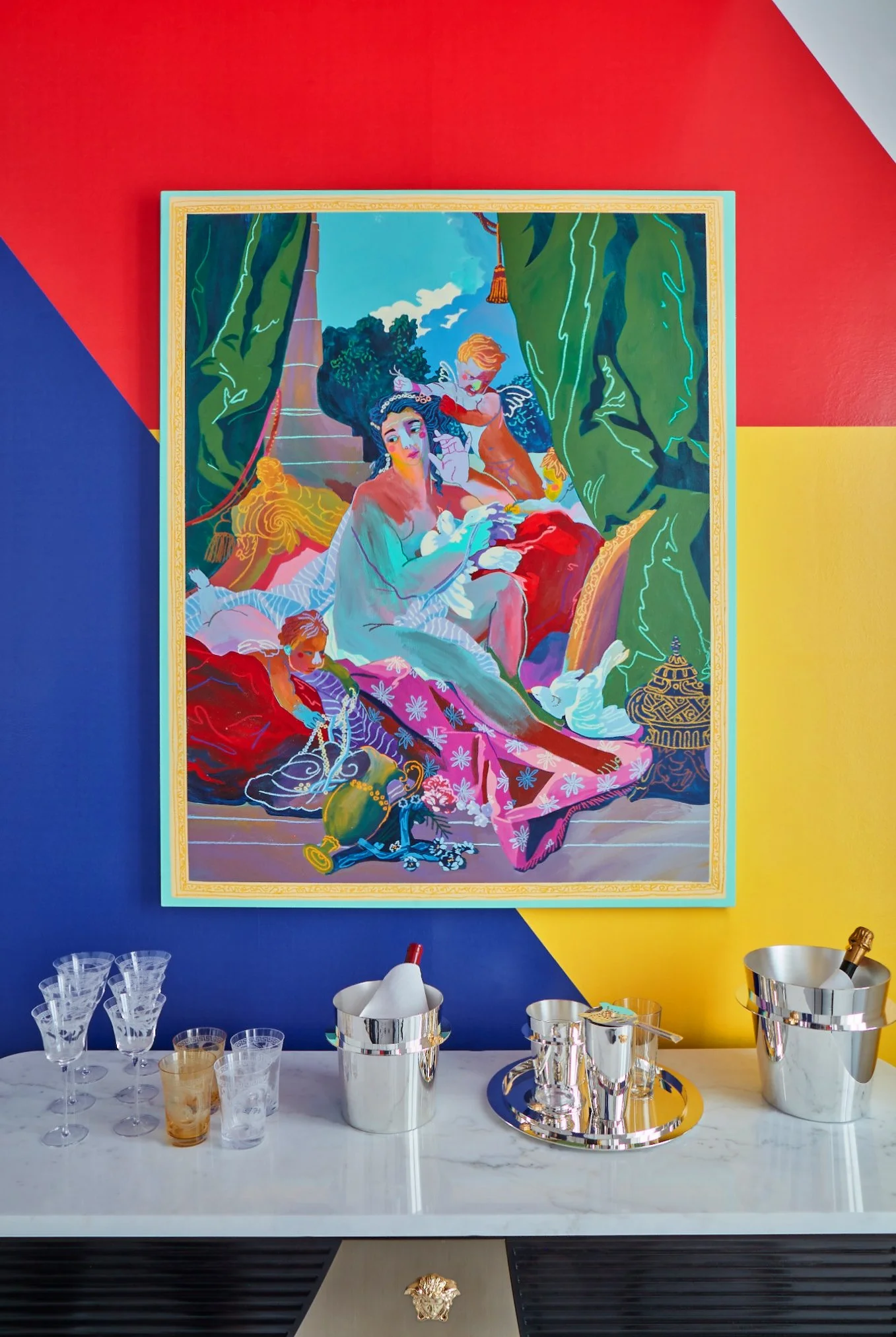 Colorful modern artwork of a woman and children on a bed, with green curtains and vibrant background, hangs on a bold red, yellow, and blue wall above a white marble countertop with champagne buckets and glasses.