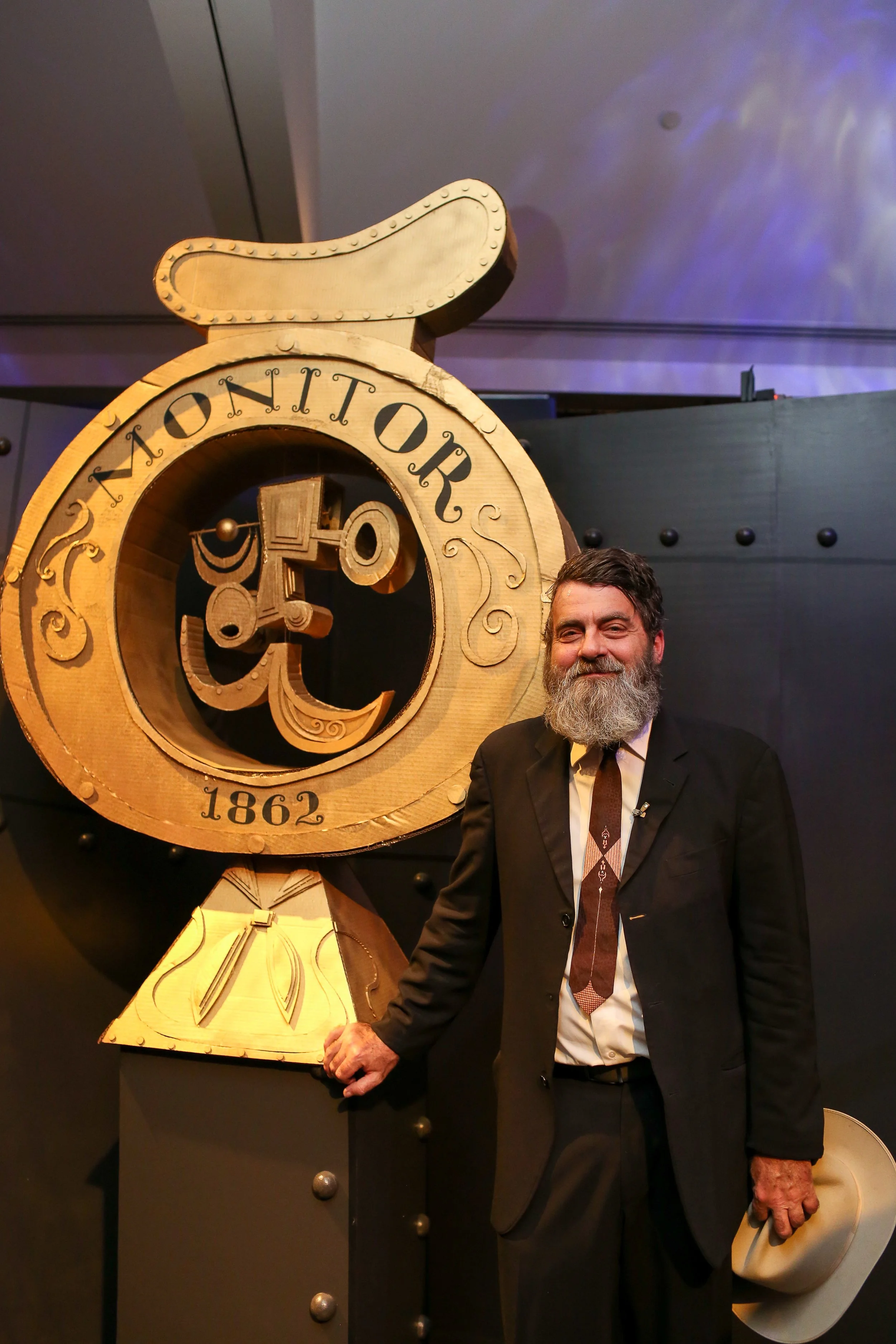 A man with a beard in a black suit standing next to a large wooden monogram clock with the words 'Montmor' and the year 1862 on it, holding a hat in his right hand.