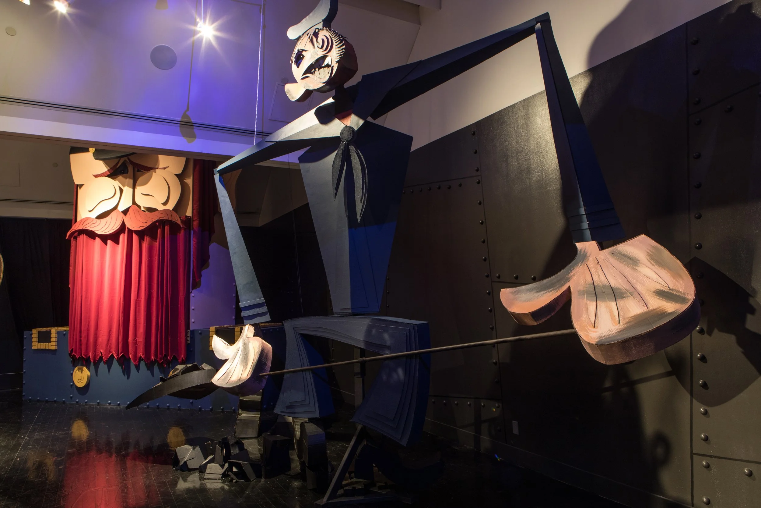 Large abstract puppet figure with exaggerated features and painted details, including a long nose, wearing a black outfit with a scarf, standing on a dark stage with a red curtain and a smaller puppet figure hanging above.