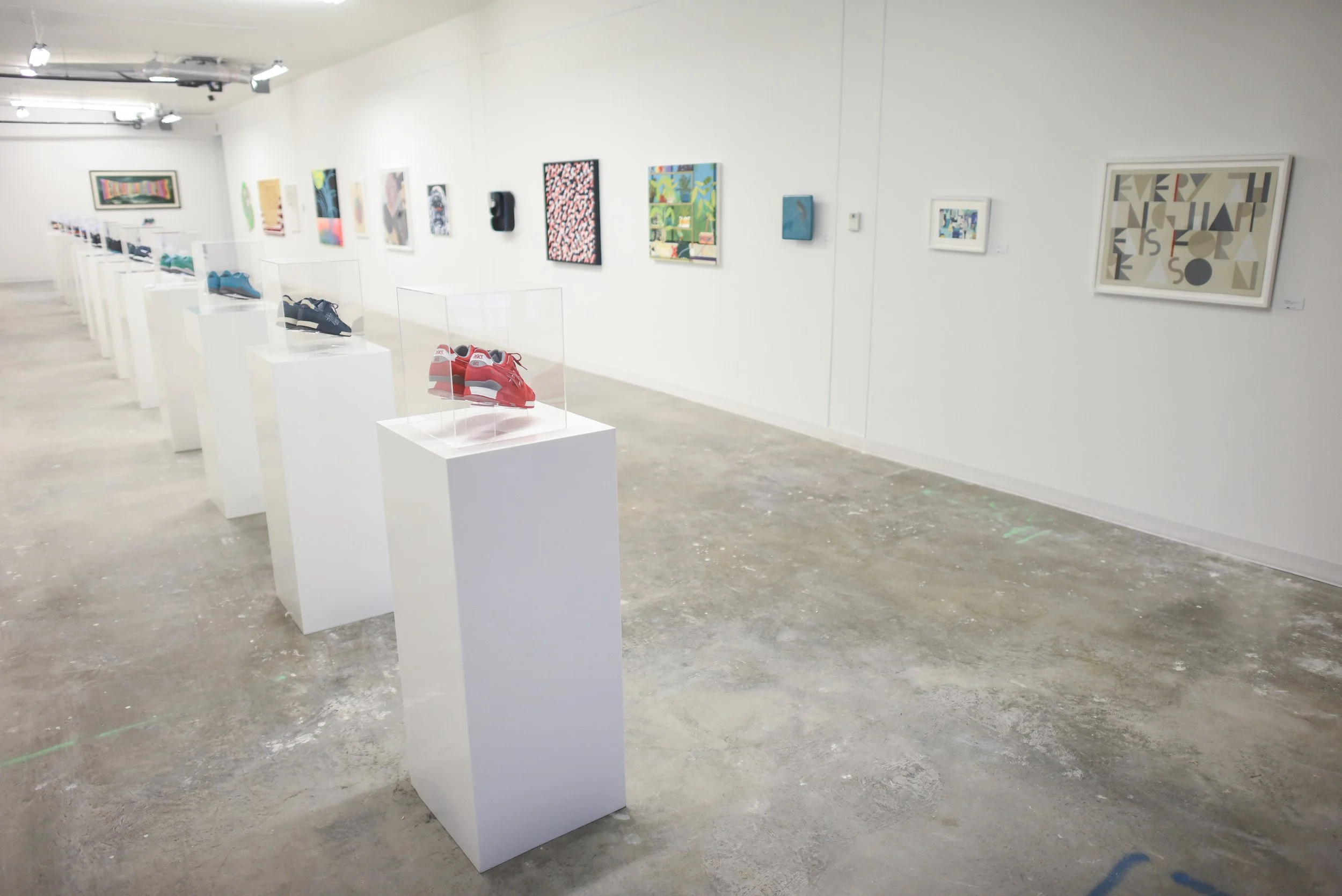 Art gallery with shoes on white pedestals and colorful artwork on white walls.