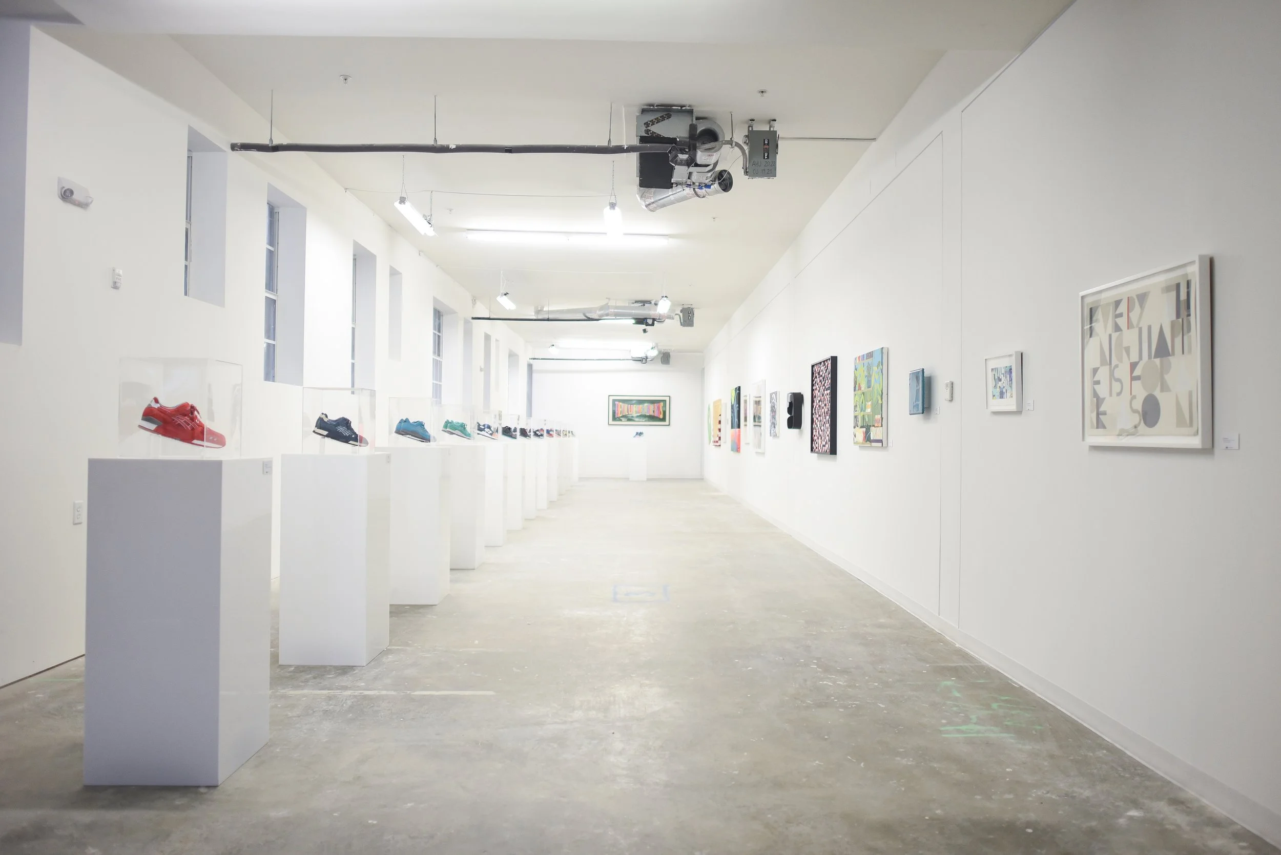 An art gallery featuring sneakers displayed on white pedestals on the left and colorful artworks on white walls on the right, with a minimalistic and well-lit interior.