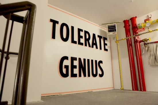 Wall with large black letters spelling 'TOLERATE GENIUS' in an industrial-style room with red pipes and yellow safety tape.