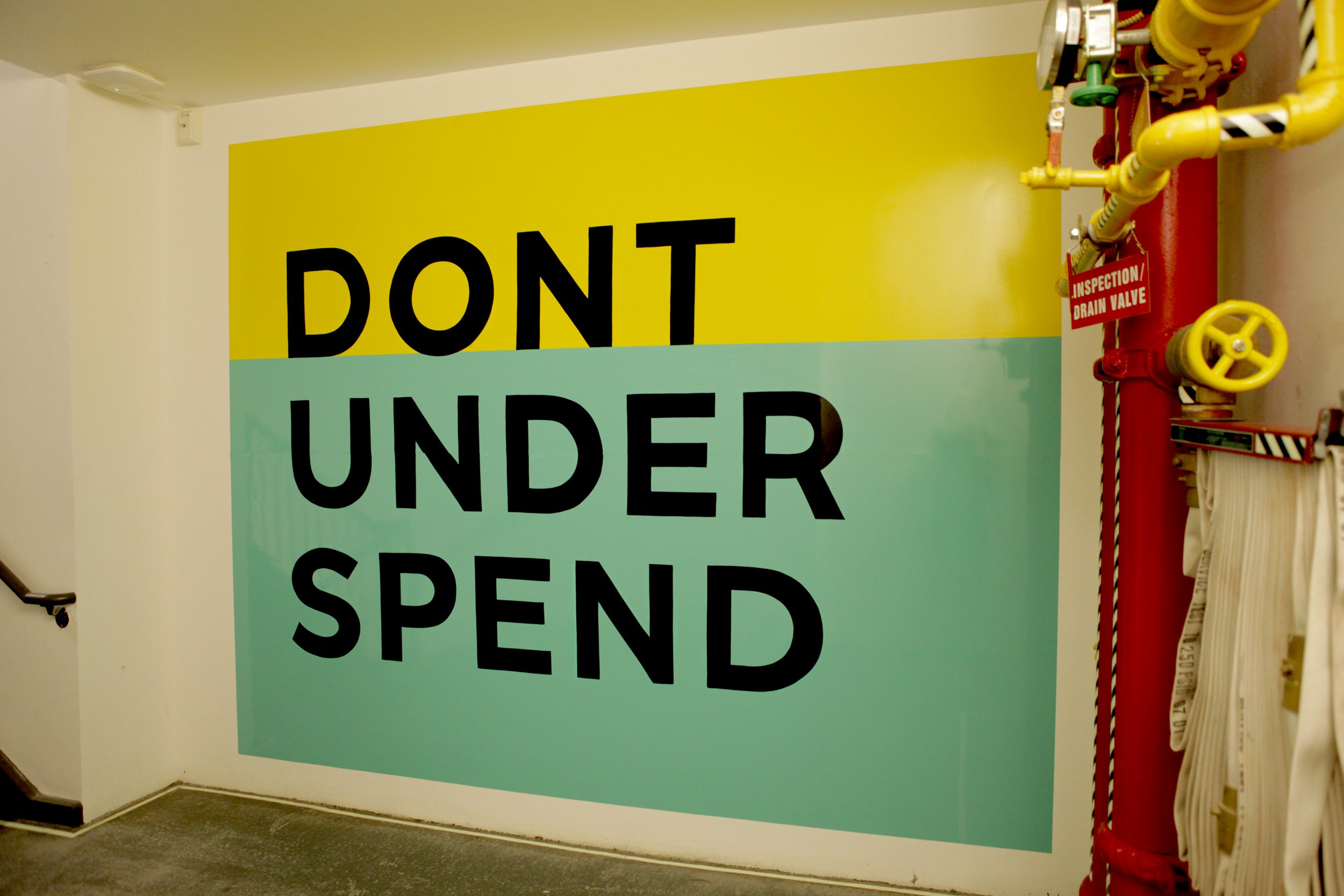 Large wall sign reading 'DON'T UNDER SPEND' with a yellow background on the top half and a light green background on the bottom half. To the right, there is a red pipe with a valve and a label that says 'inspection drain valve'.