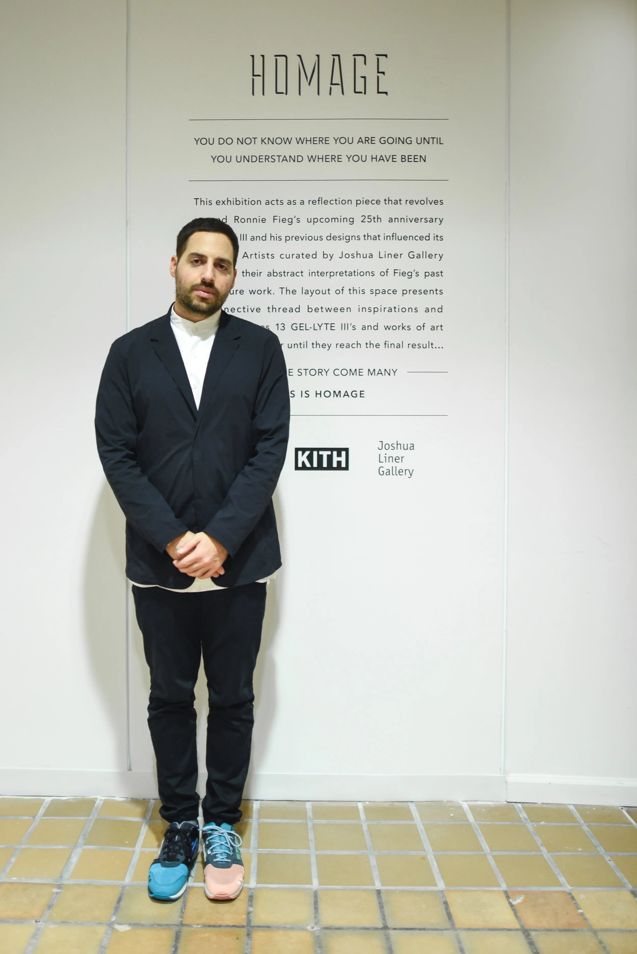 A man standing indoors against a white wall with a poster, wearing a dark jacket, white shirt, dark pants, and colorful sneakers.