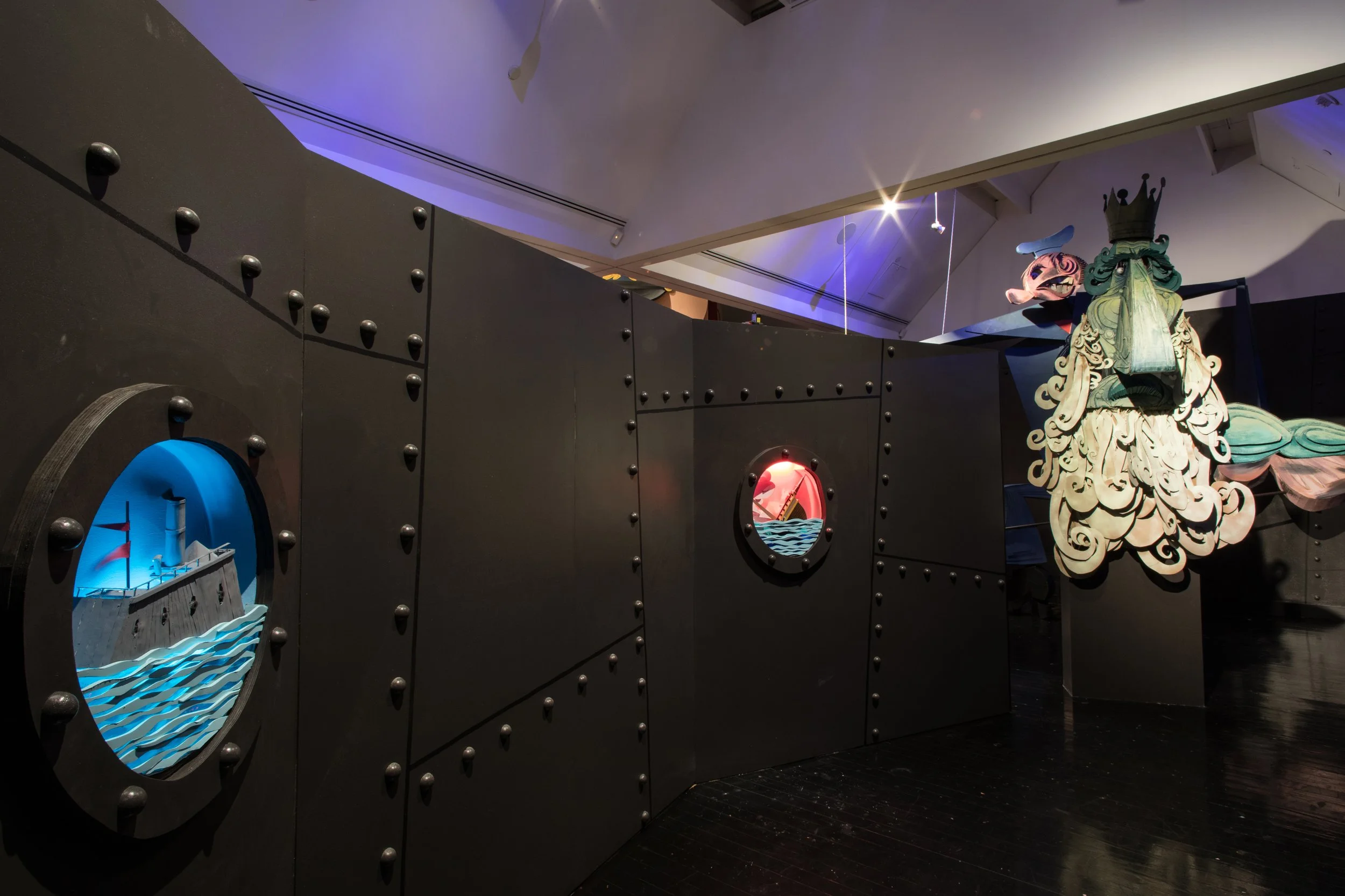 Decorative black wall with portholes showing scenes of a battleship, a ship at harbor, and a mythical creature with a crown and flowing hair