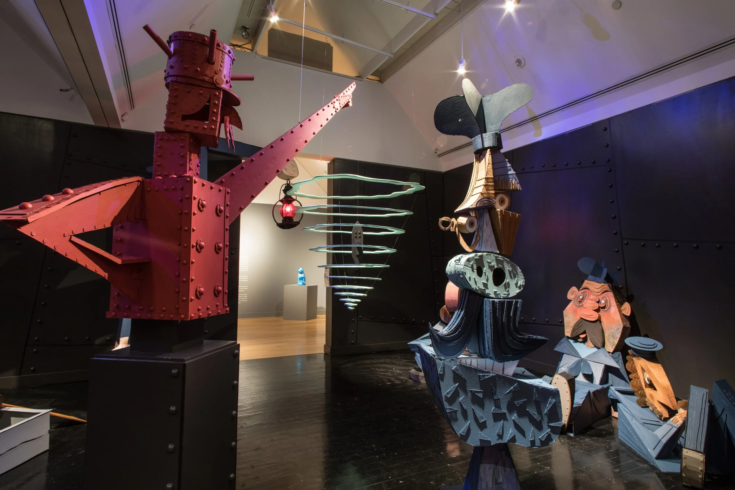 Artistic sculptures of abstract faces and objects in a gallery with black walls and wooden floors, illuminated by spotlights.