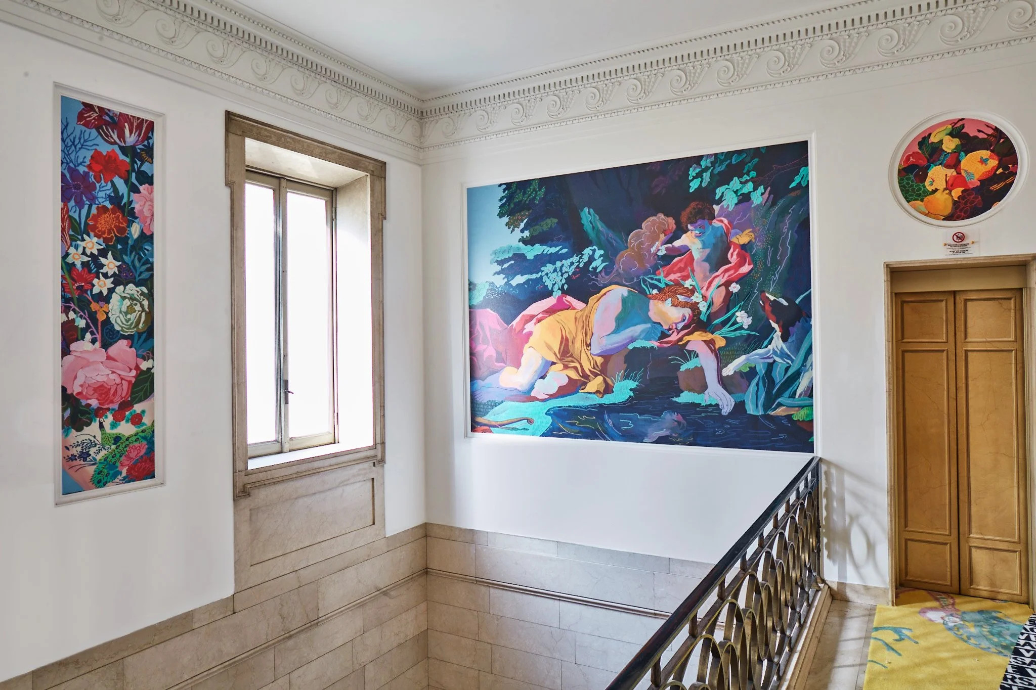 Gallery interior with colorful paintings on white walls, a window, a wooden door, and a decorative railing.