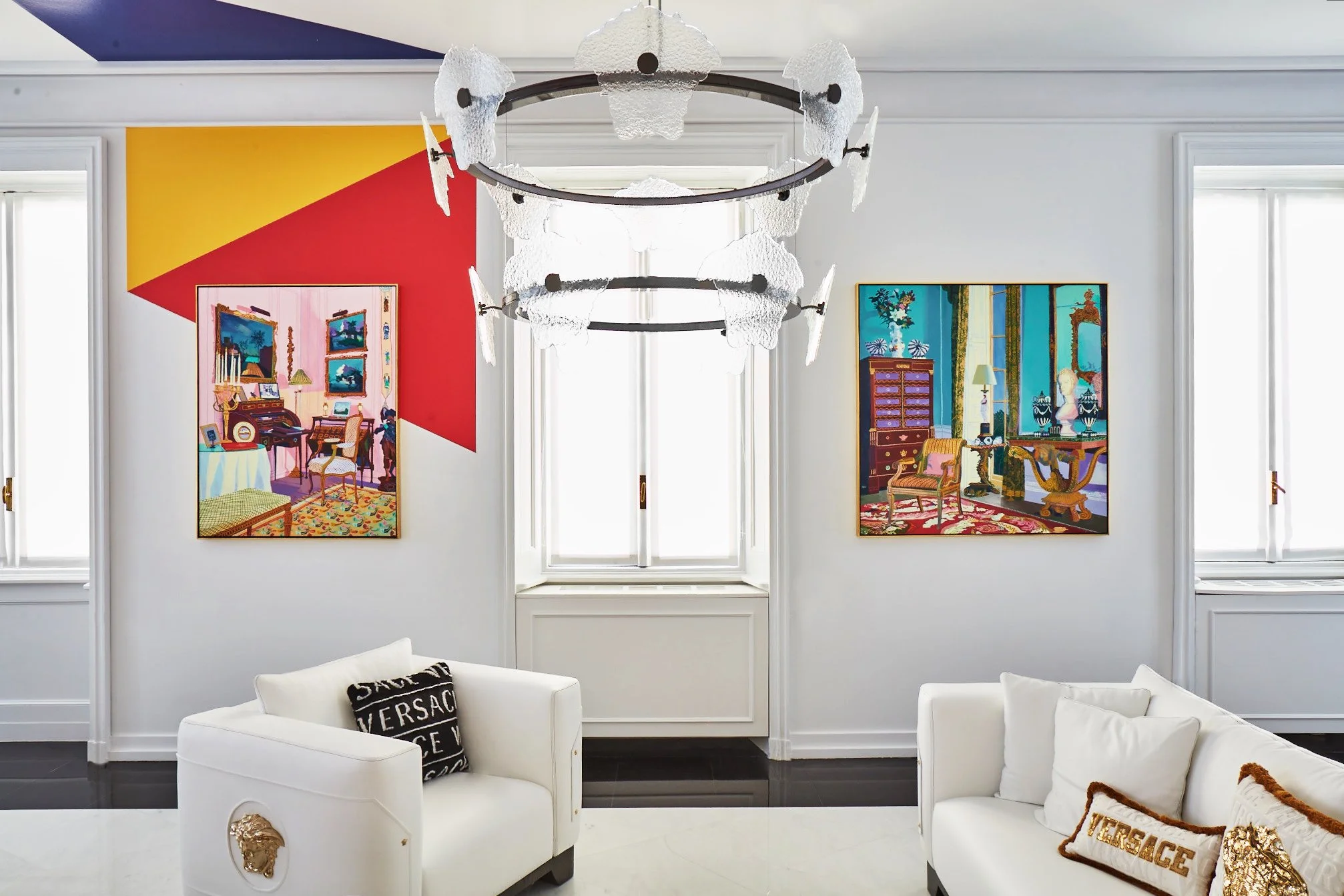 A modern living room with white walls, three large windows, and a contemporary chandelier. Two colorful paintings hang on the wall, one on each side of a central window. White couches with luxury brand pillows are in the foreground.