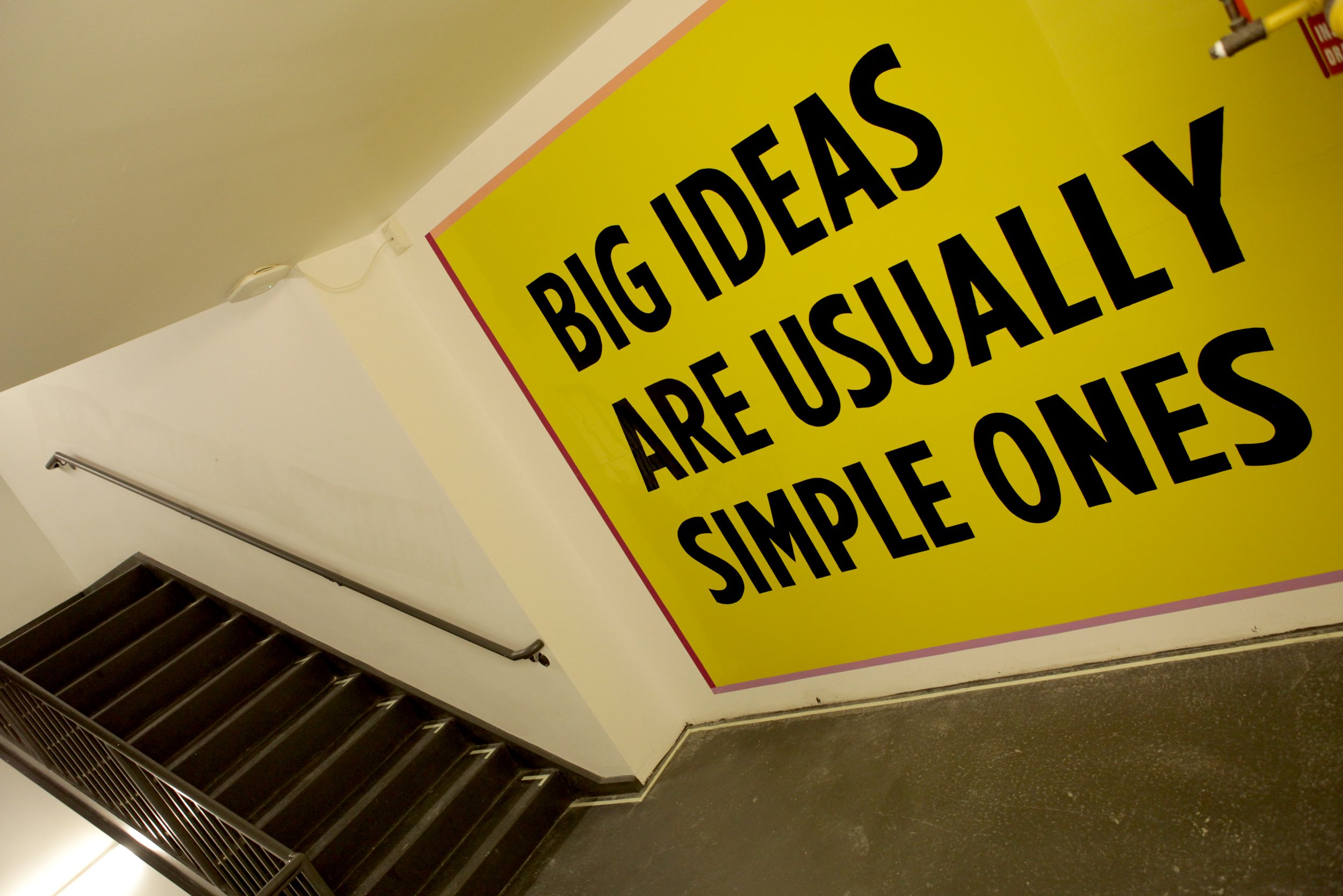 Large yellow sign on wall reading 'Big ideas are usually simple ones' near staircase in building.