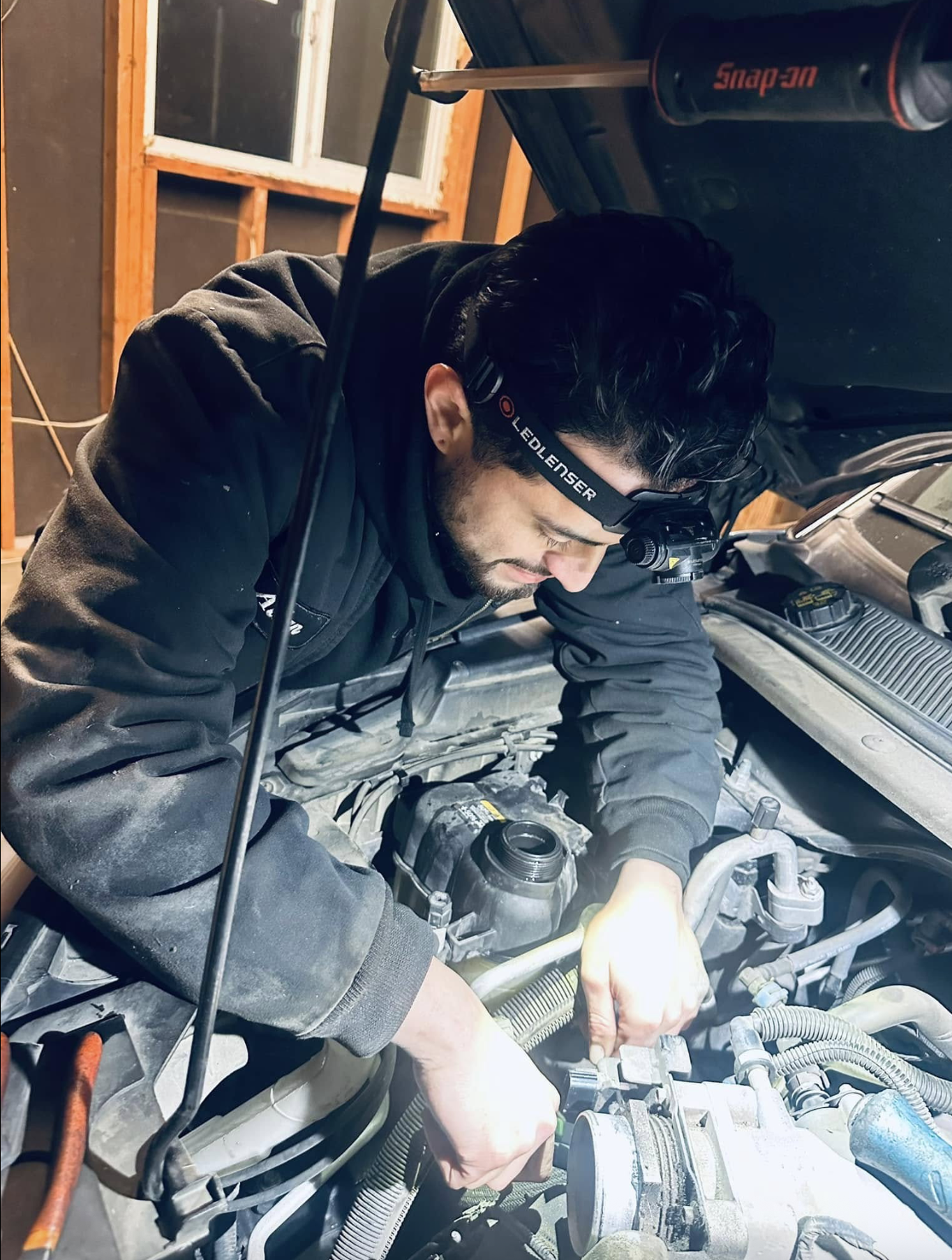 A man working on a car engine in a garage, wearing a headlamp and black hoodie, leaning over the engine with tools in hand.