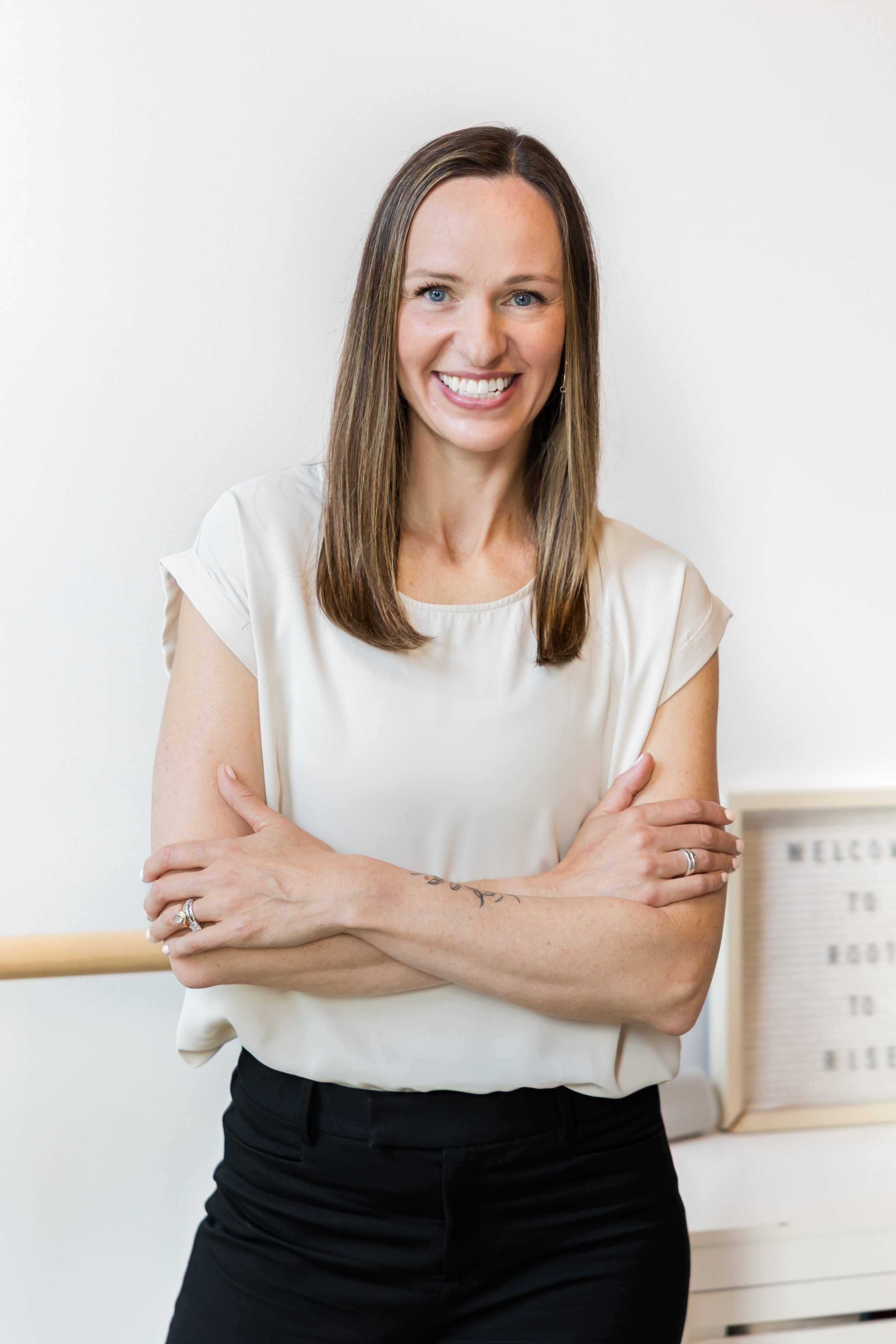 A smiling woman with long brown hair, wearing a white top and black pants, standing with arms crossed in front of a white wall and a sign that says 'Welcome to...'