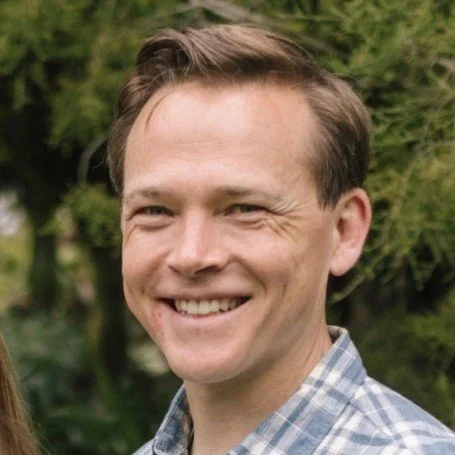 A smiling man with short brown hair wearing a blue plaid shirt outdoors with trees in the background.