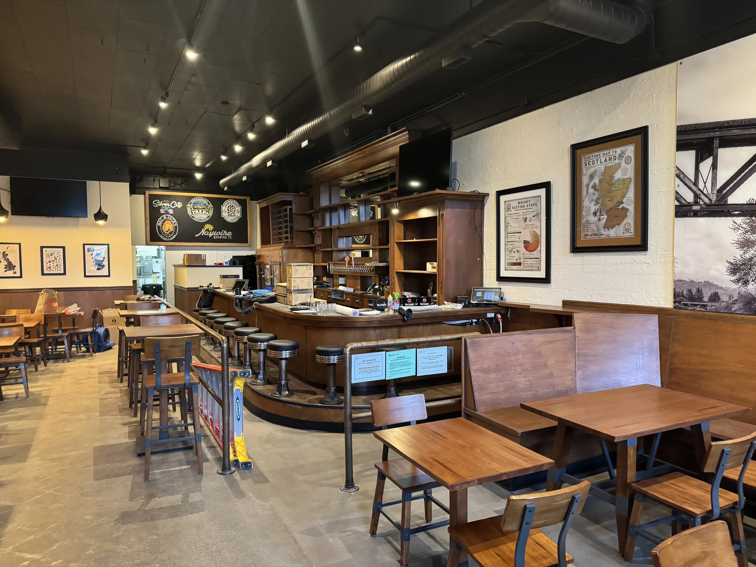Interior of a restaurant or bar with wooden tables and chairs, a curved wooden bar counter with bar stools, framed posters on the wall, and a ceiling with black paint and exposed ductwork.
