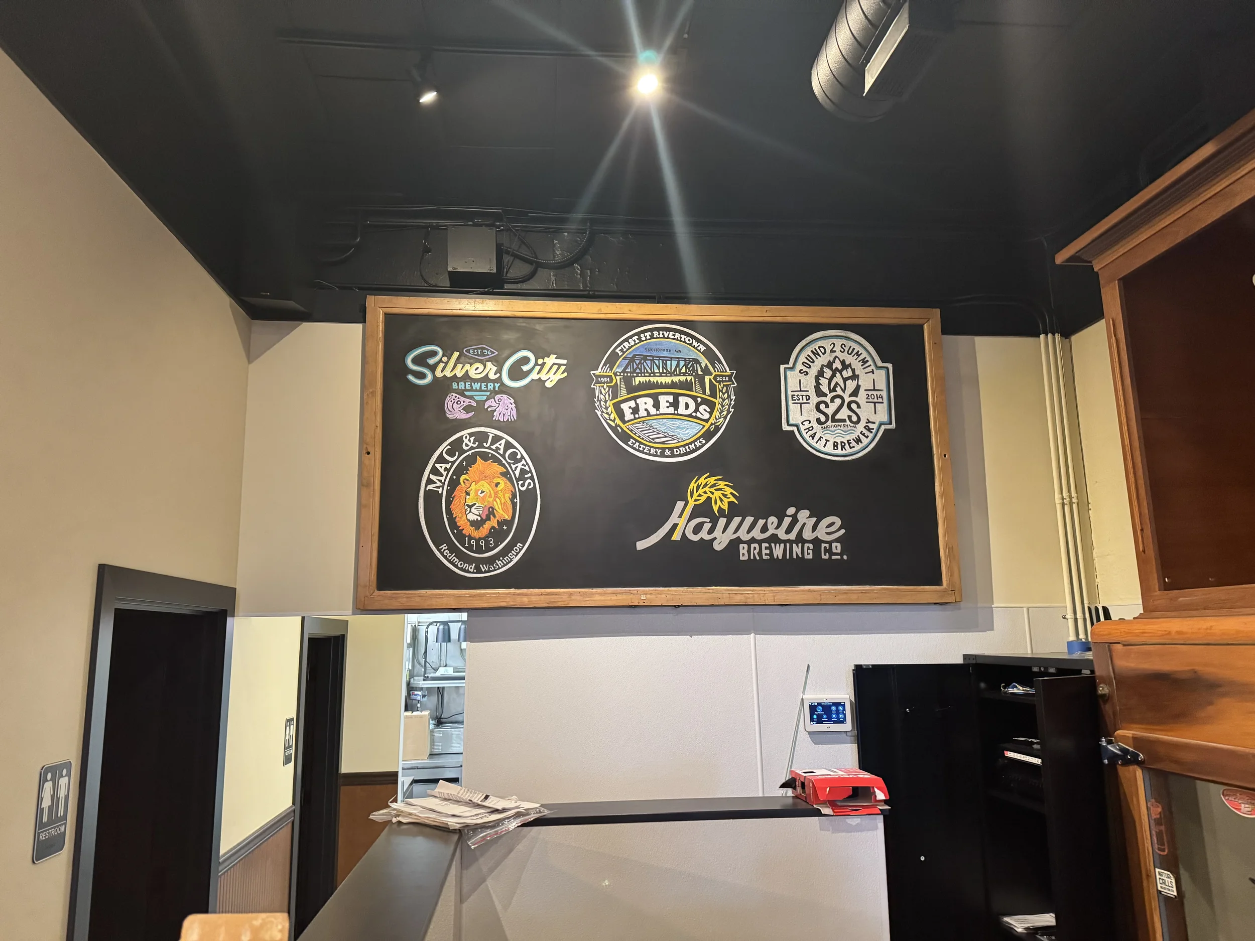 A wall with a large blackboard displaying logos of various breweries, including Silver City Brewery, FRED'S, S2S Craft Brewery, Mac & Jack's, and Haywire Brewing Co., located inside a brewery or taproom, with a partial view of a counter and a restroo