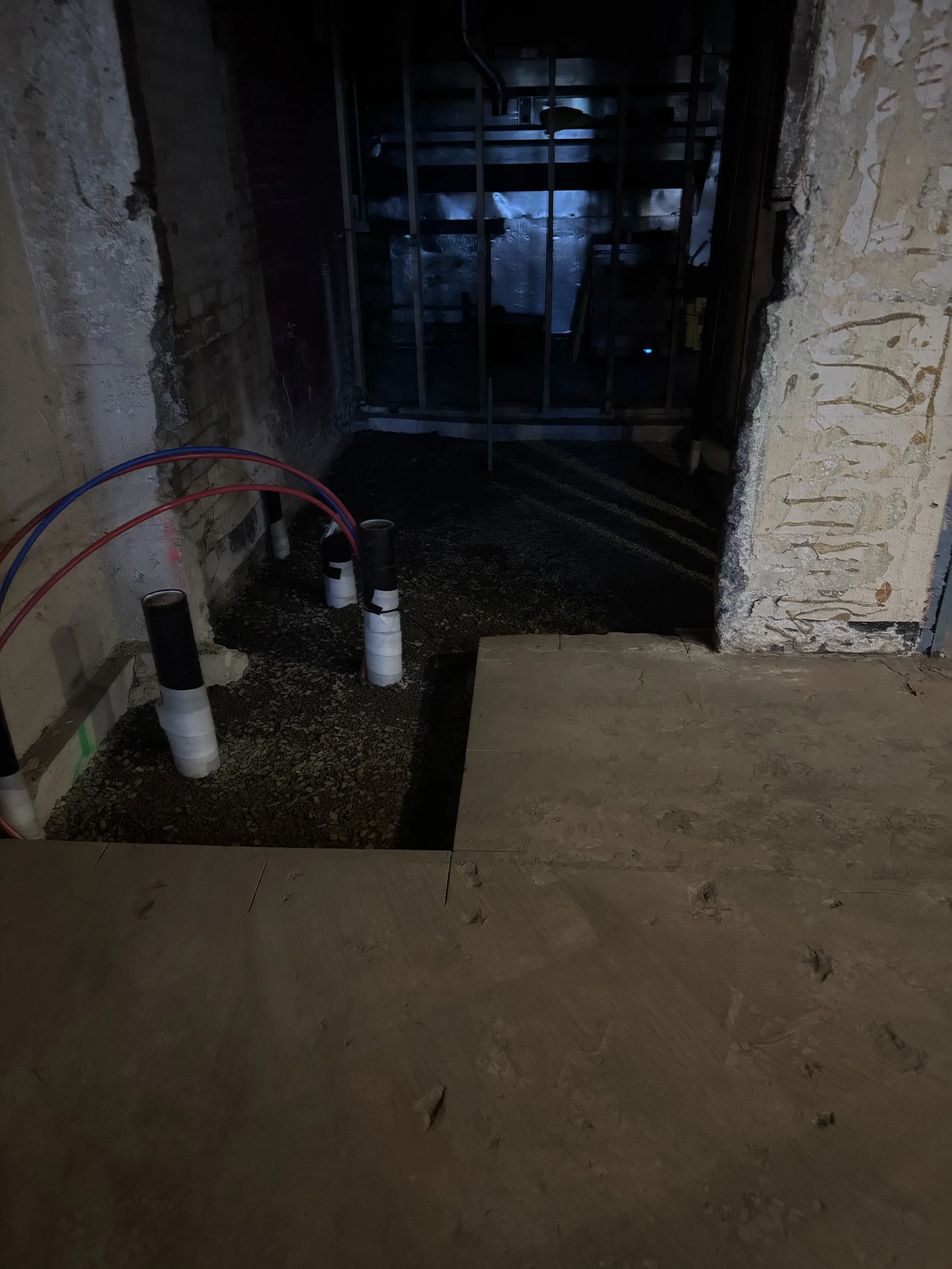 Interior of a construction area showing exposed plumbing pipes and an unfinished wall with framing. Some flooring is partially installed, with dirt and gravel visible on the ground.
