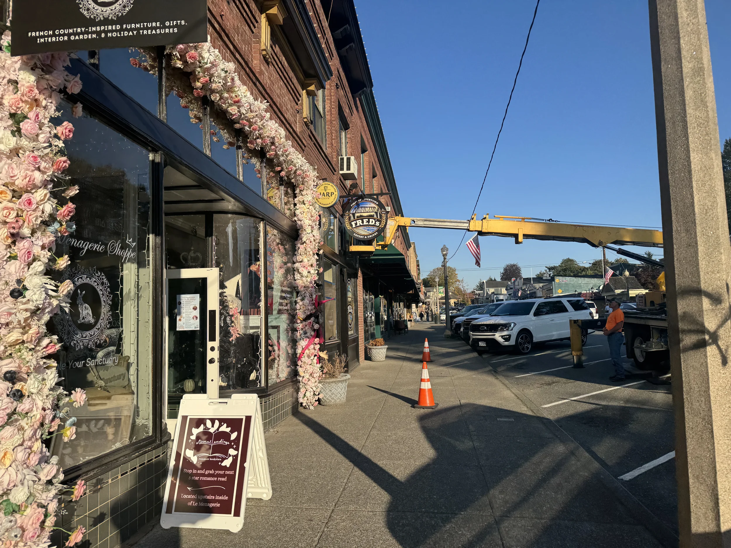 Street view showing storefront decorated with pink and white flower garland, sidewalk with traffic cones, parked cars, and a construction worker near a utility pole, with a clear blue sky overhead.