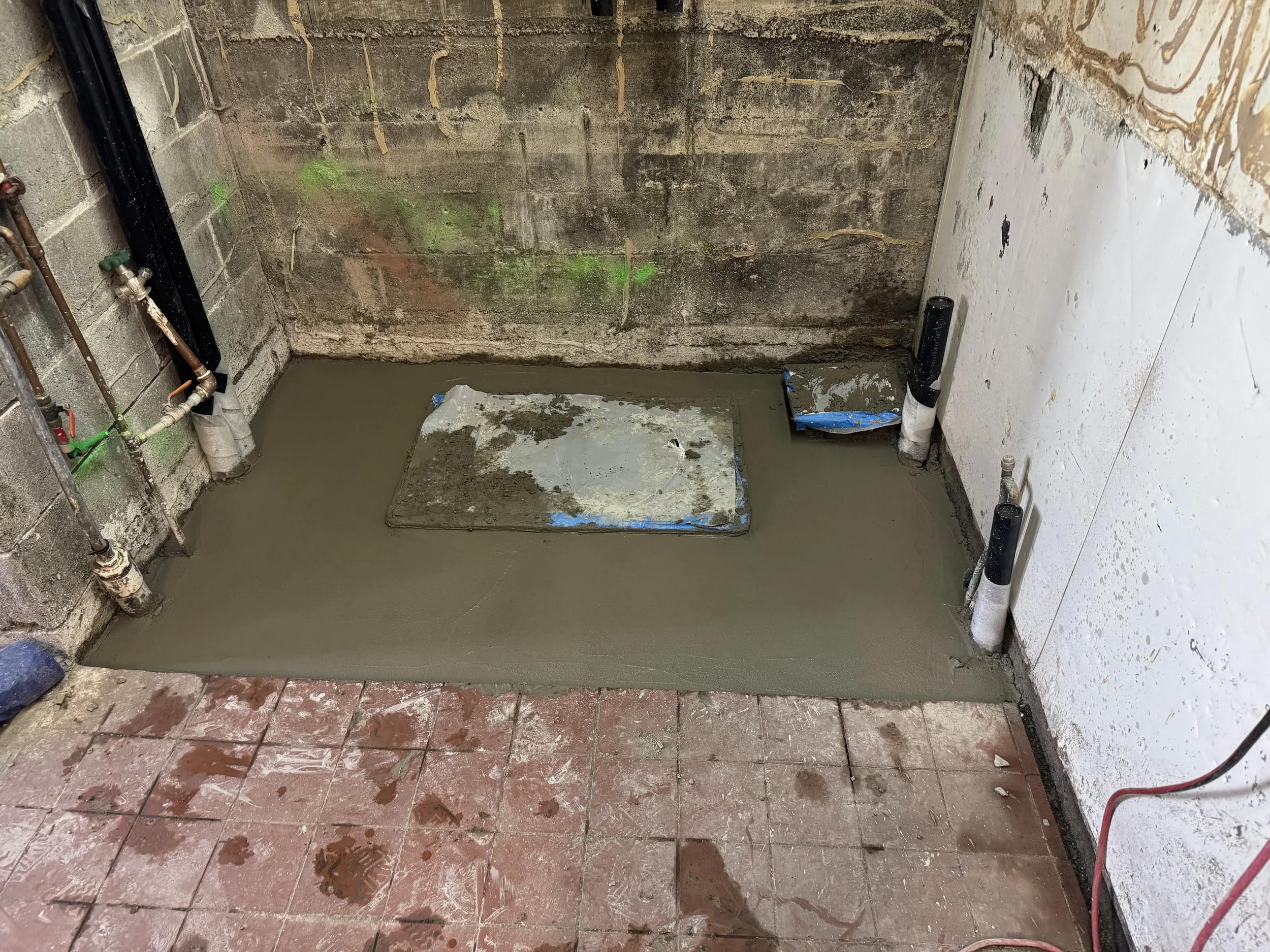A construction or renovation site showing a partially finished bathroom with concrete and pipes. The floor has exposed bricks and wet cement, and there is a metal plate in the center. Green and black water pipes are visible along the walls, and there