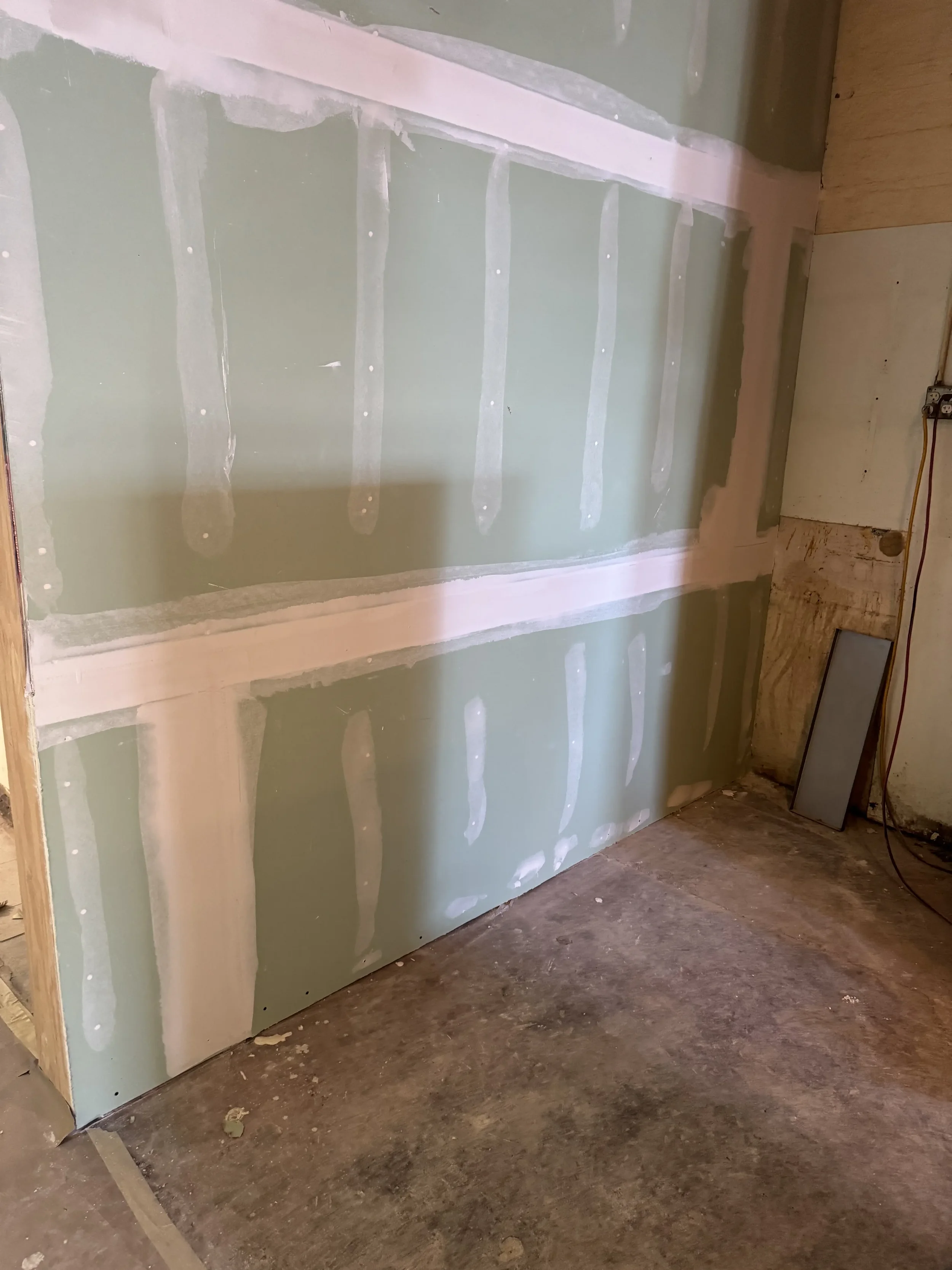 Green drywall with white joint compound and tape during construction or renovation, with a concrete floor and unfinished wall edges.