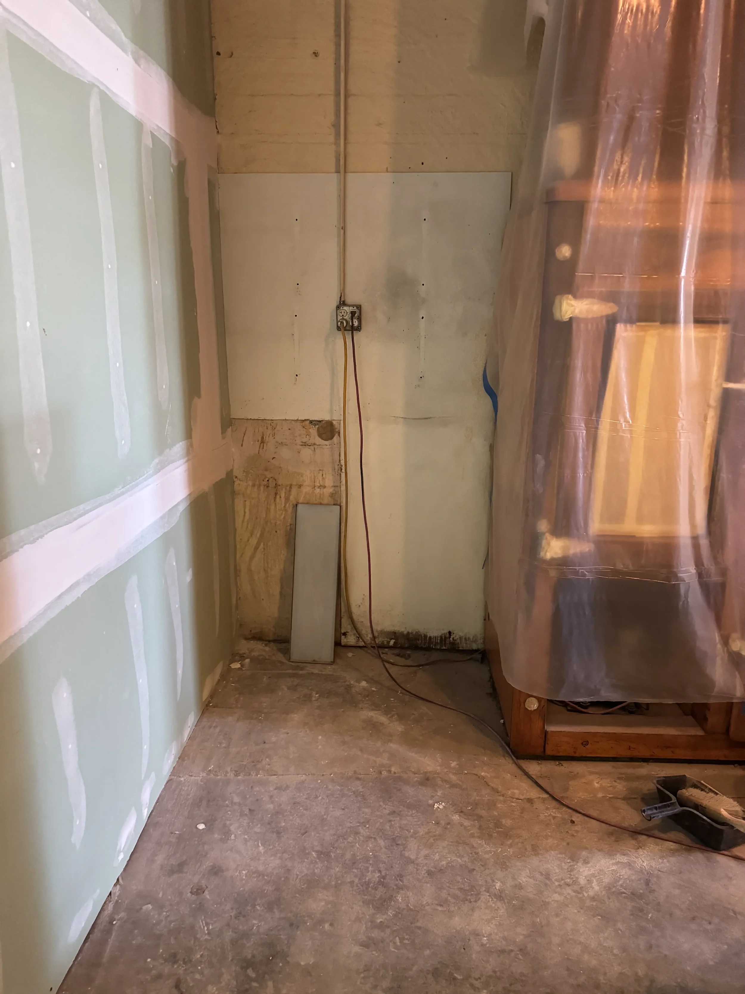 Empty room with drywall and a concrete floor, with an electrical outlet on the wall and a wire hanging down, partially covered with plastic sheeting.