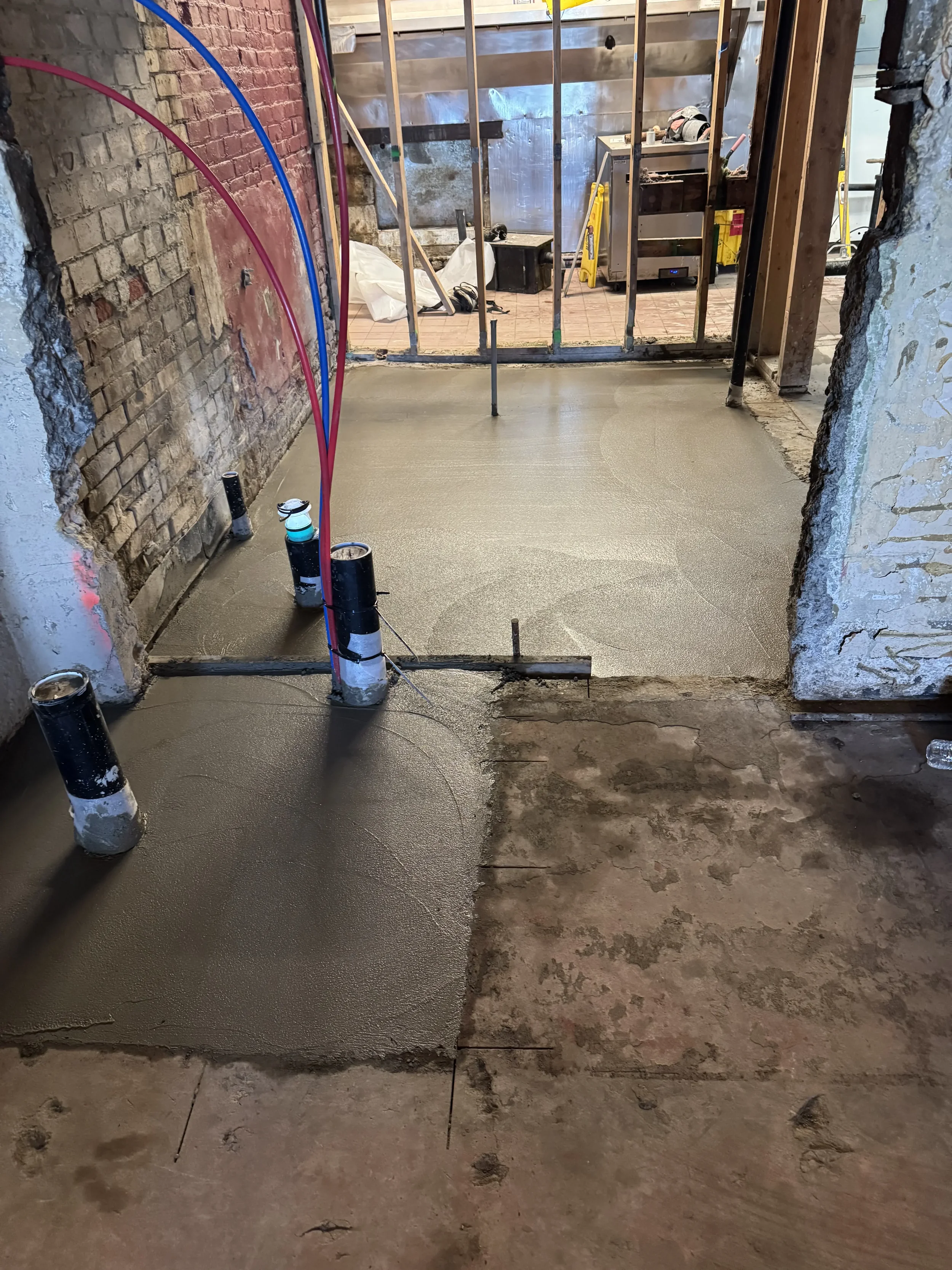Construction site with freshly poured concrete on the floor, partially cured, with exposed brick walls and construction materials and equipment in the background.