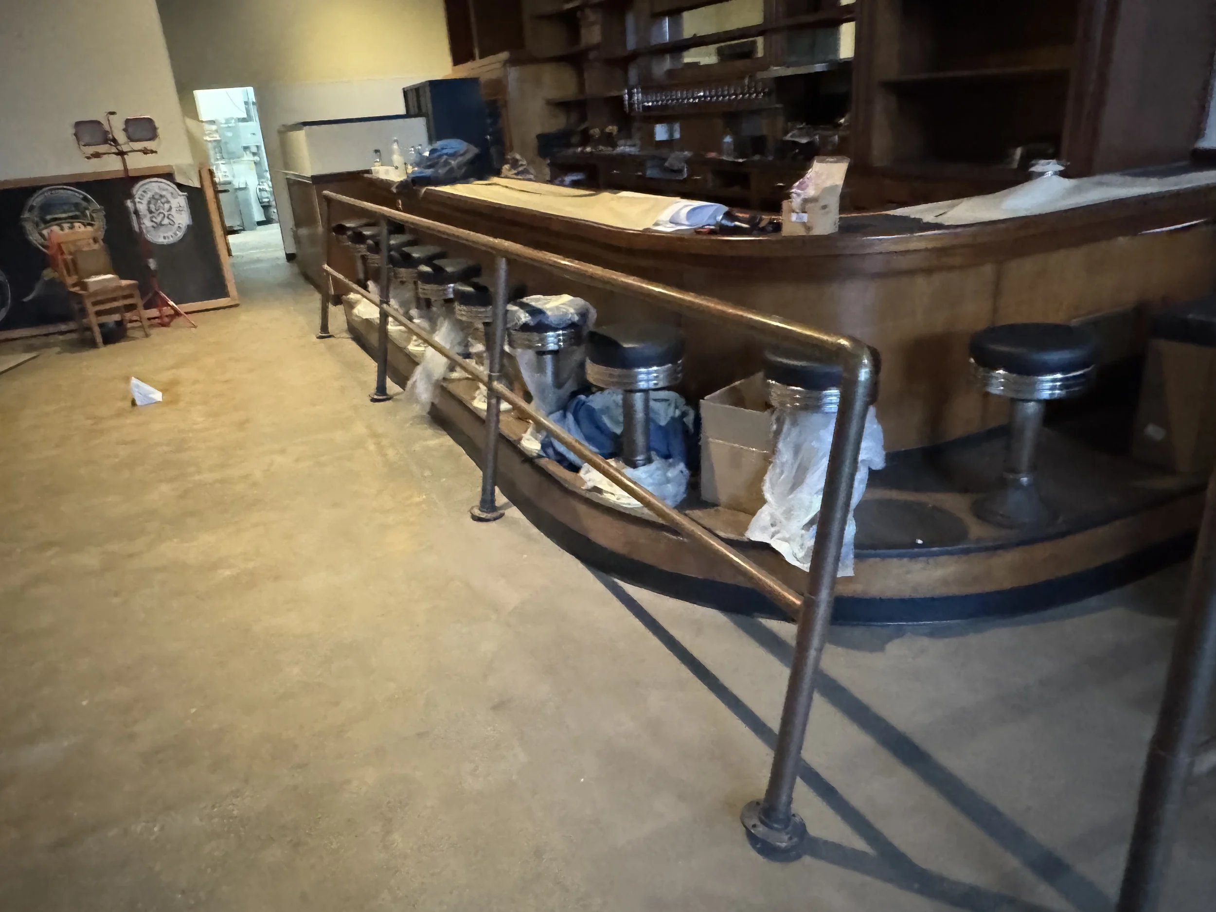 Empty bar counter with bar stools, some supplies and boxes scattered, in a dimly lit, possibly closed restaurant or pub.