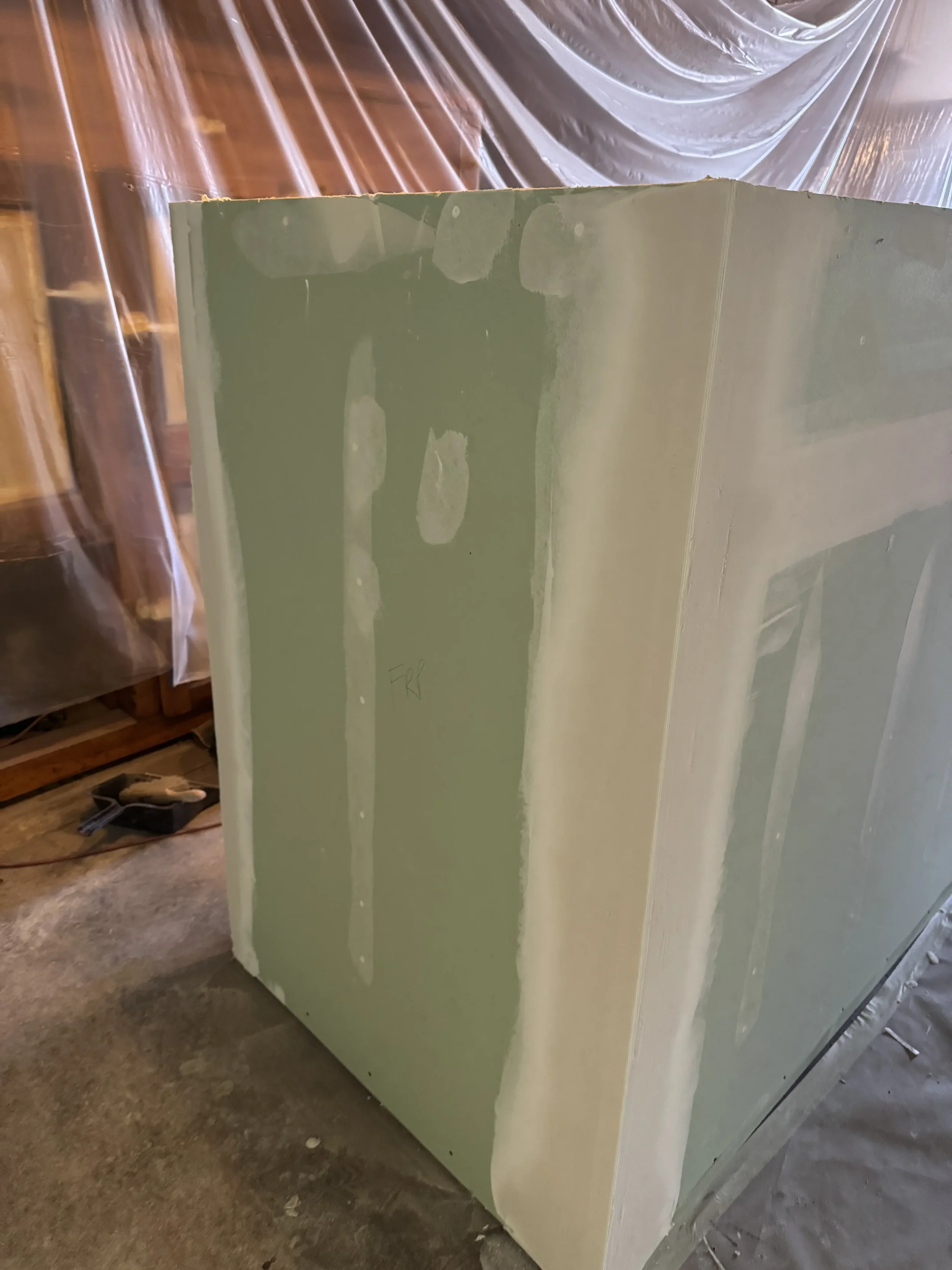 A large drywall panel with visible joint compound applied along the seams, standing on a concrete floor in a room under renovation.