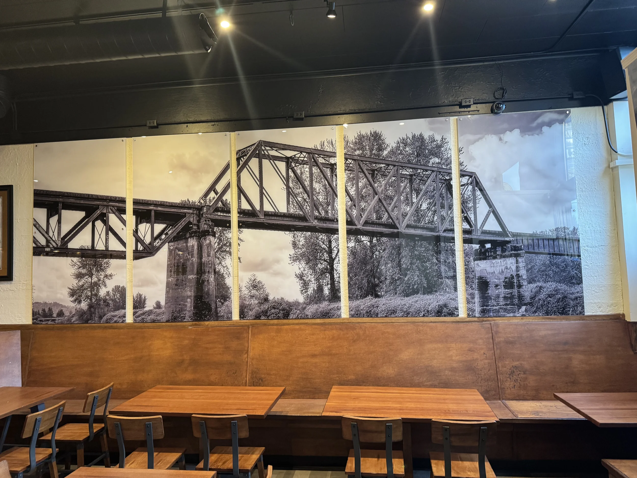 A black-and-white mural of a bridge with trees and sky in the background, mounted on a wall in a cafe or restaurant setting.