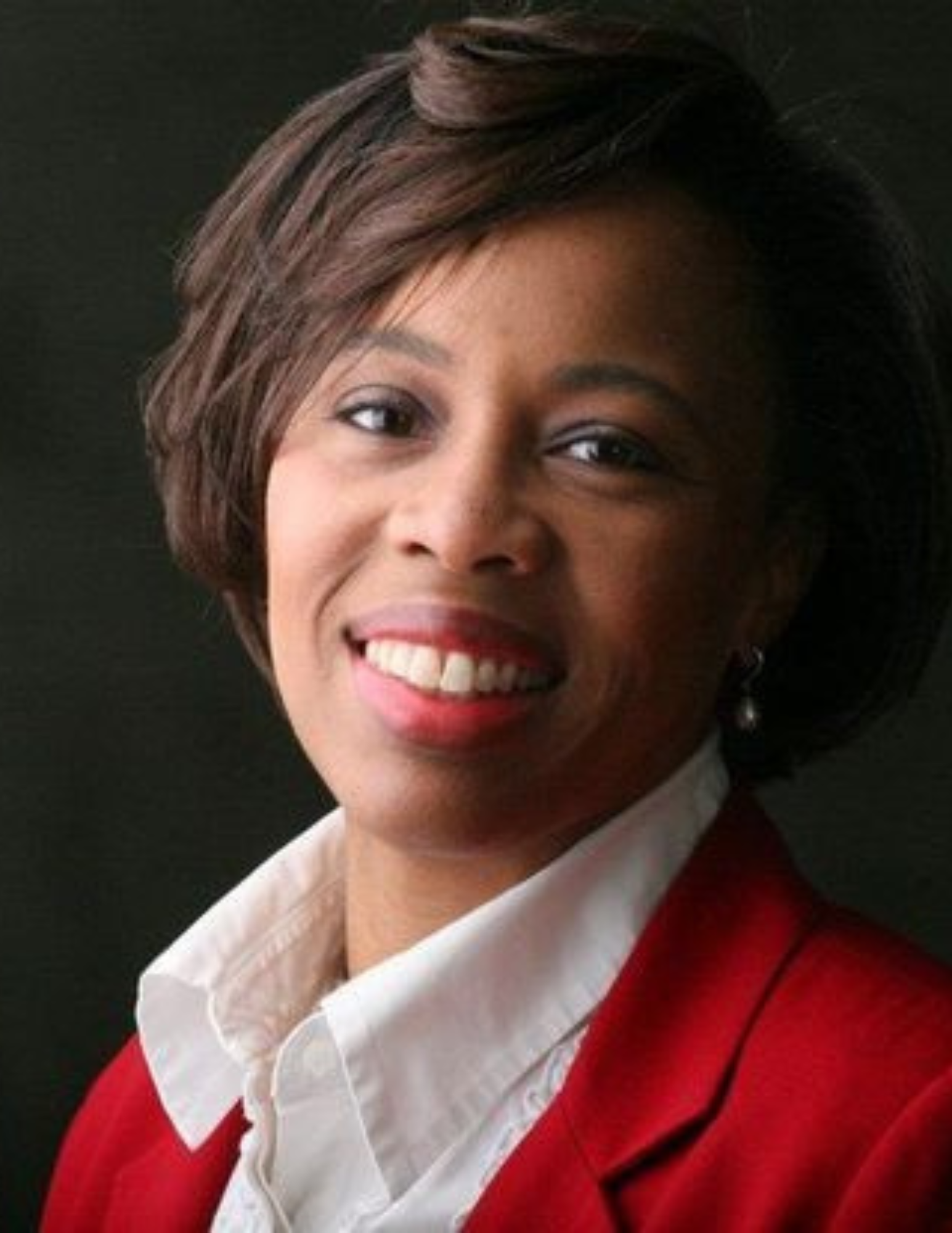 Portrait of a woman with short, styled hair, wearing a white collared shirt and a red blazer, smiling against a dark background.
